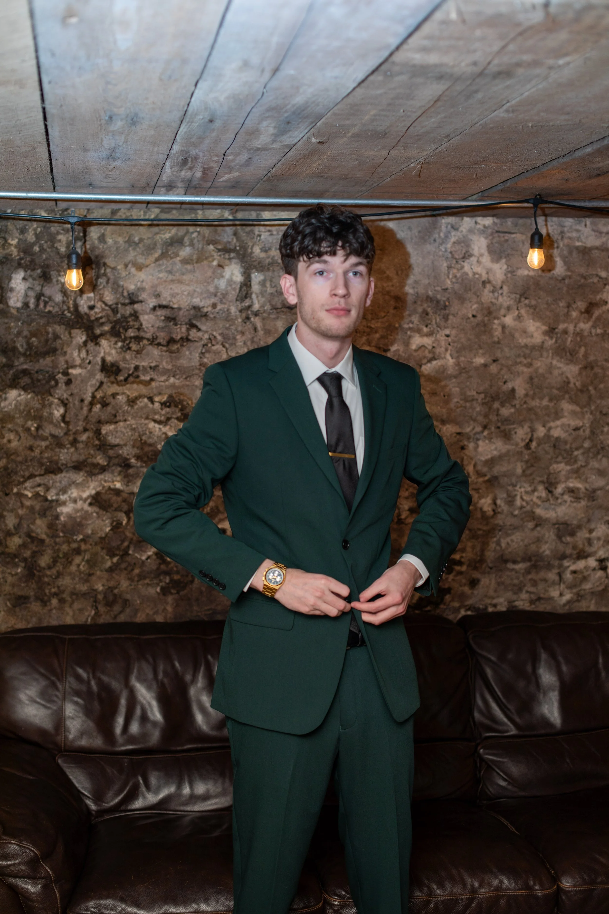 A young man in a green suit, white shirt, and black tie standing in front of a stone wall with string lights hanging above, adjusting his jacket.