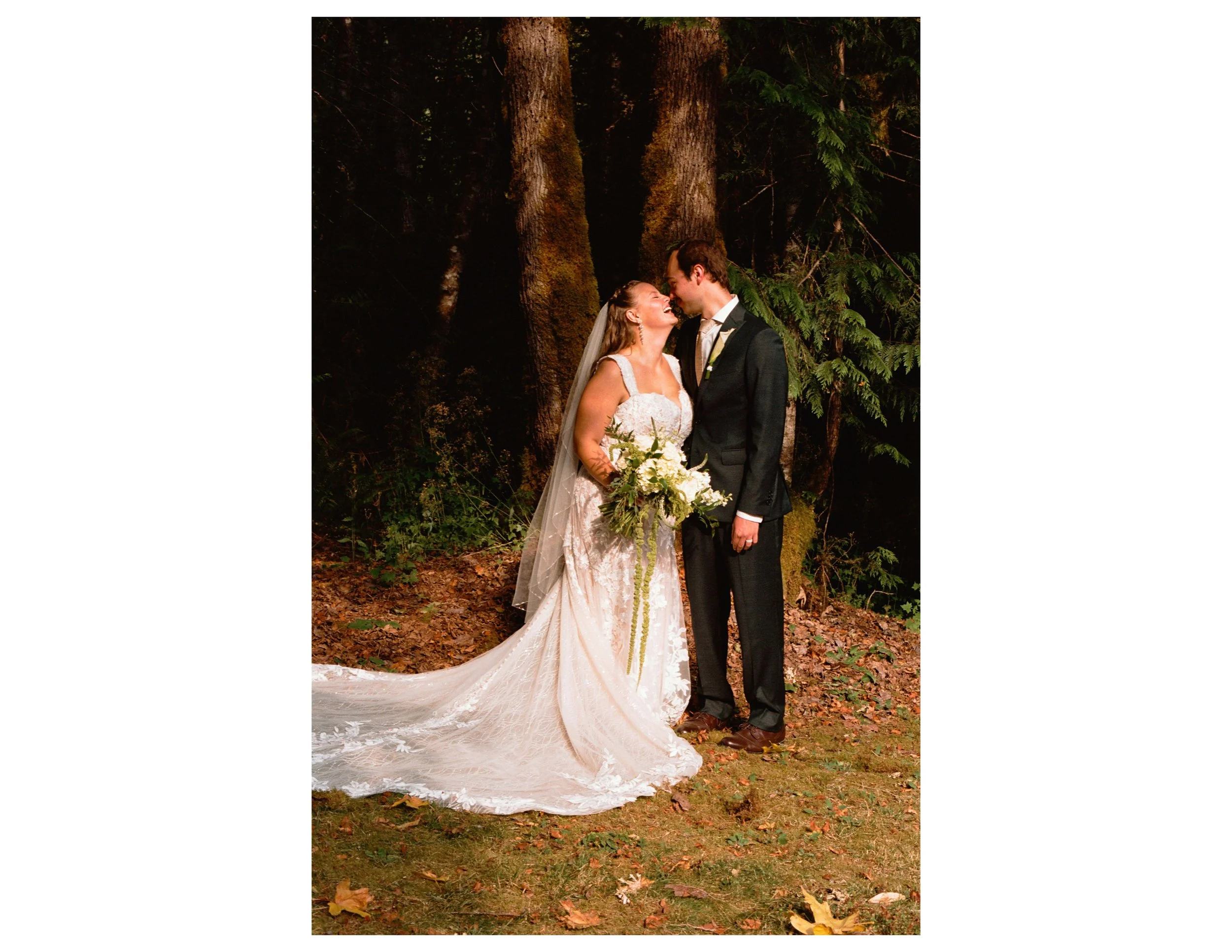 A bride and groom stand close together outdoors at night in front of tall trees, sharing a joyful moment. The bride wears a white lace wedding gown with a long train and holds a bouquet of white flowers. The groom is in a dark suit with a white shirt