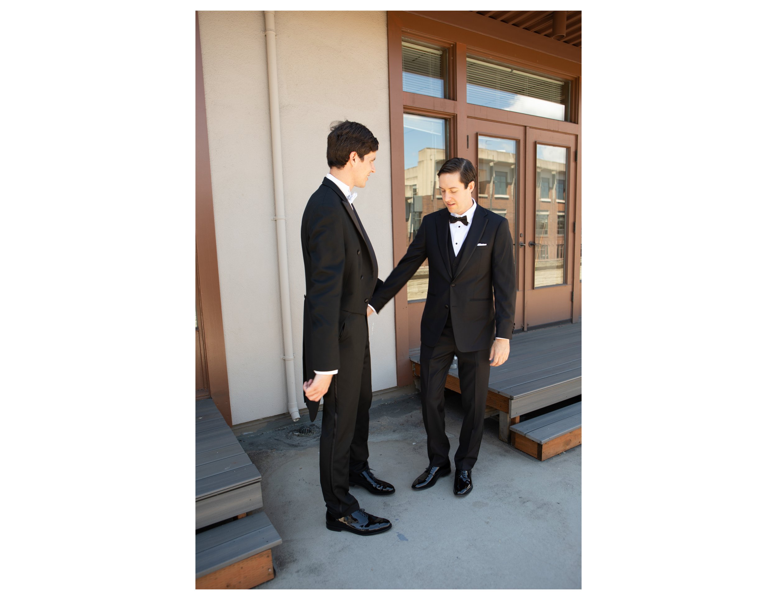 Two men dressed in black tuxedos shake hands outdoors on a balcony or patio with wooden flooring, against a building with large windows and a beige wall.