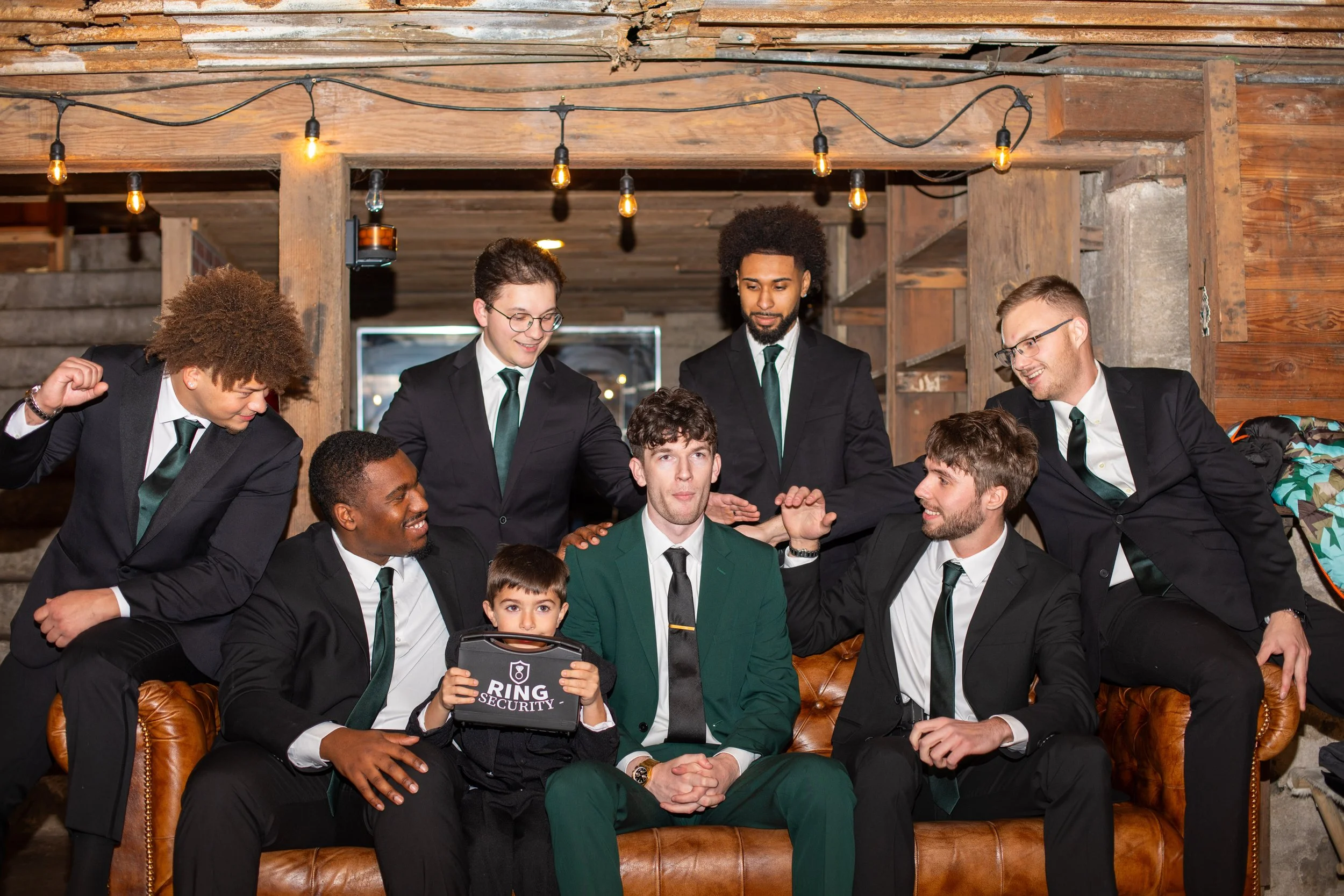 Group of young men in suits gathered around and sitting on a leather couch, one holding a toy briefcase that says 'Ring Security,' in a rustic indoor setting with wooden walls and string lights.