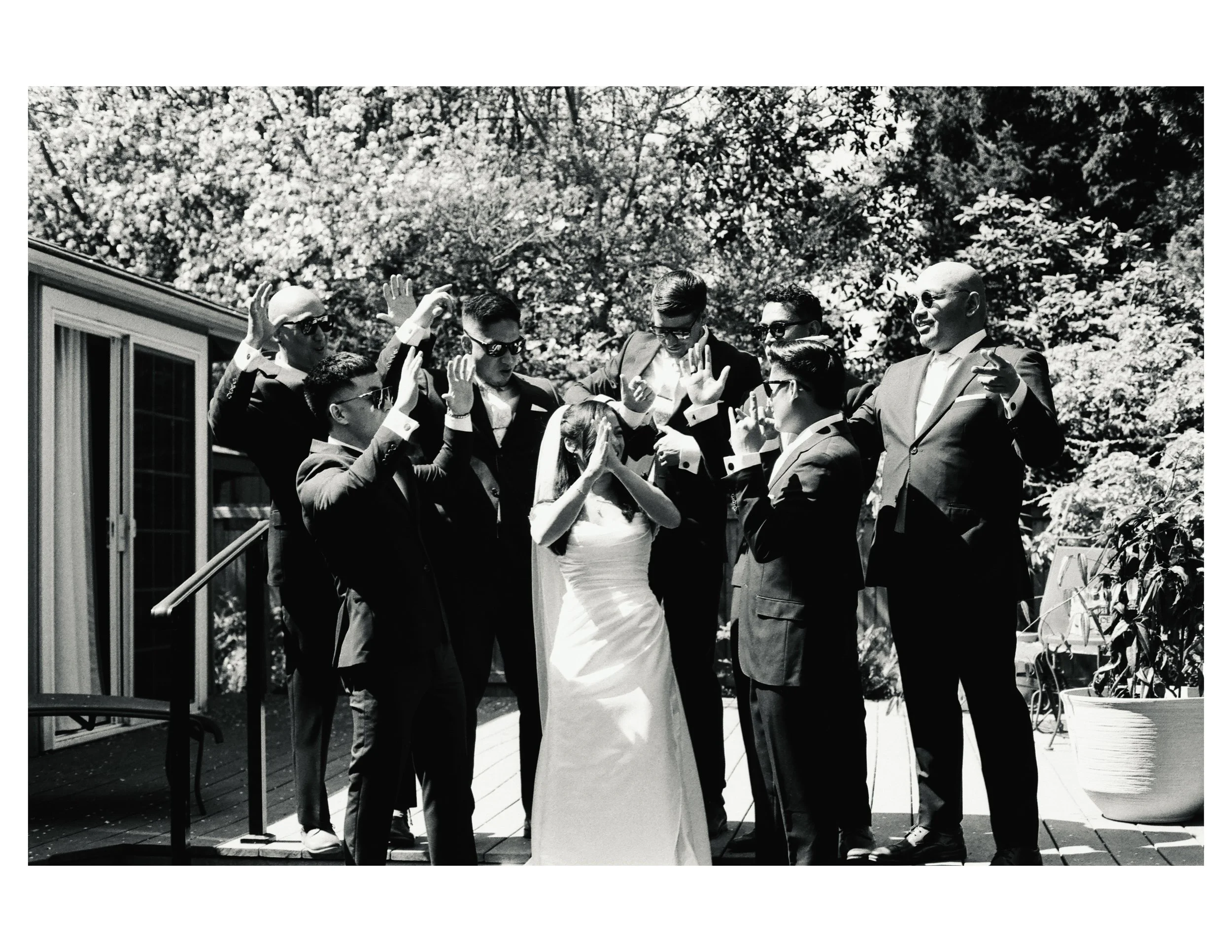 A group of people dressed in formal attire, possibly at a wedding reception, engaging in conversations and gestures outdoors, with trees and a building in the background.