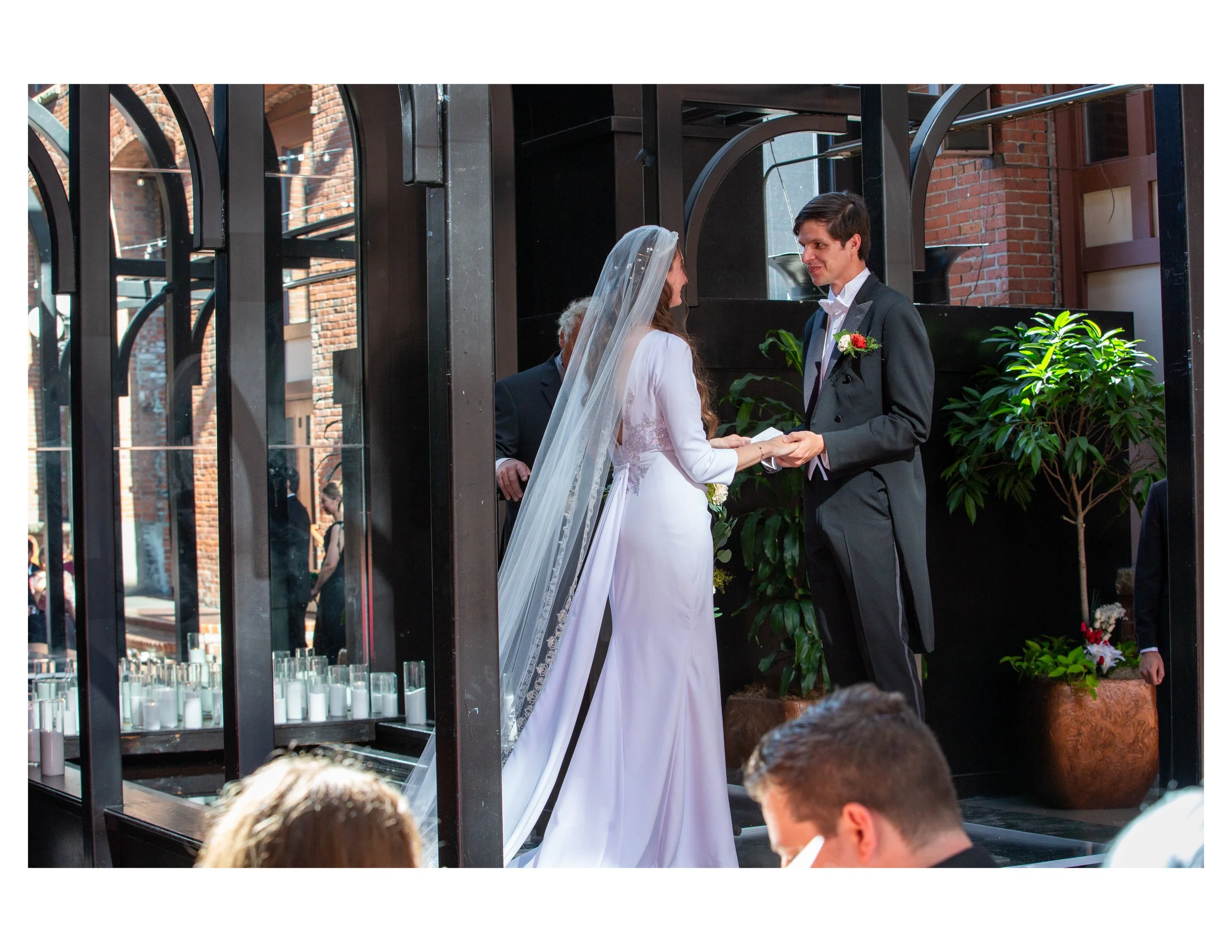 A bride and groom exchanging vows inside a glass-walled venue with brick walls and greenery.