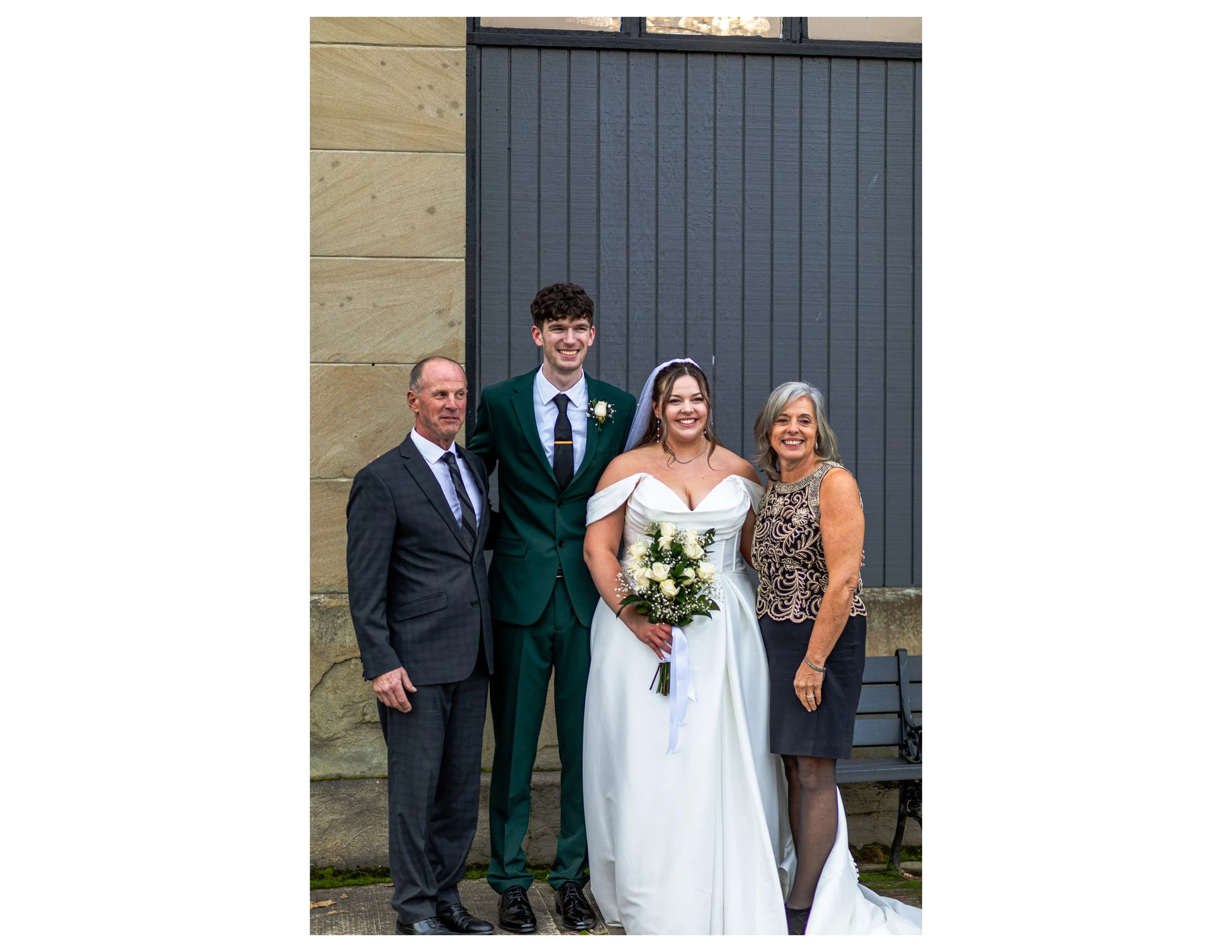 A group of five people, including a bride and groom, standing outside near a wall with a large dark gray door and a beige stucco section, smiling at the camera.