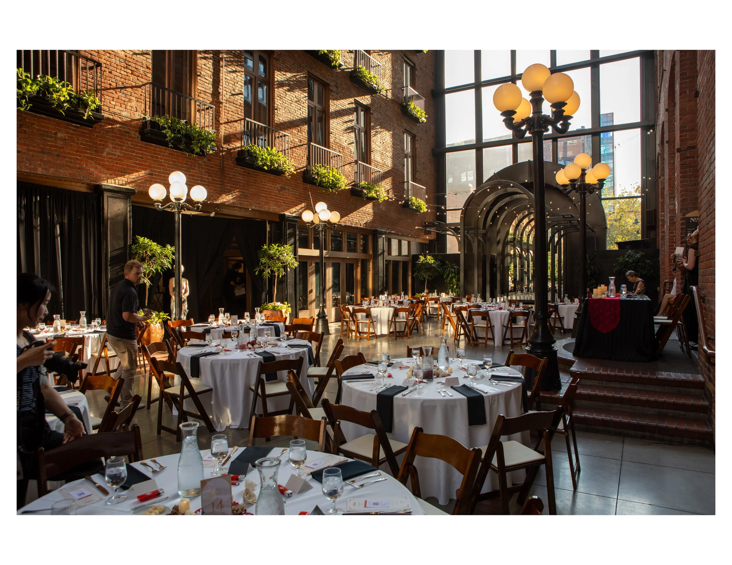 Interior of a restaurant with round tables covered in white tablecloths, set with glasses, utensils, and napkins, featuring a brick wall, large windows, potted plants, and decorative lighting fixtures.