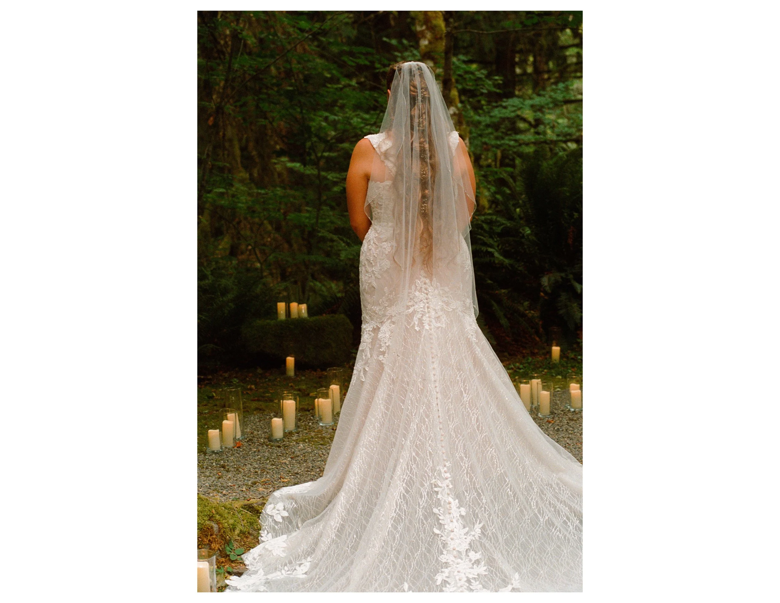 A woman in a lace wedding dress with a long train and a veil standing in a forest, surrounded by candles on the ground.