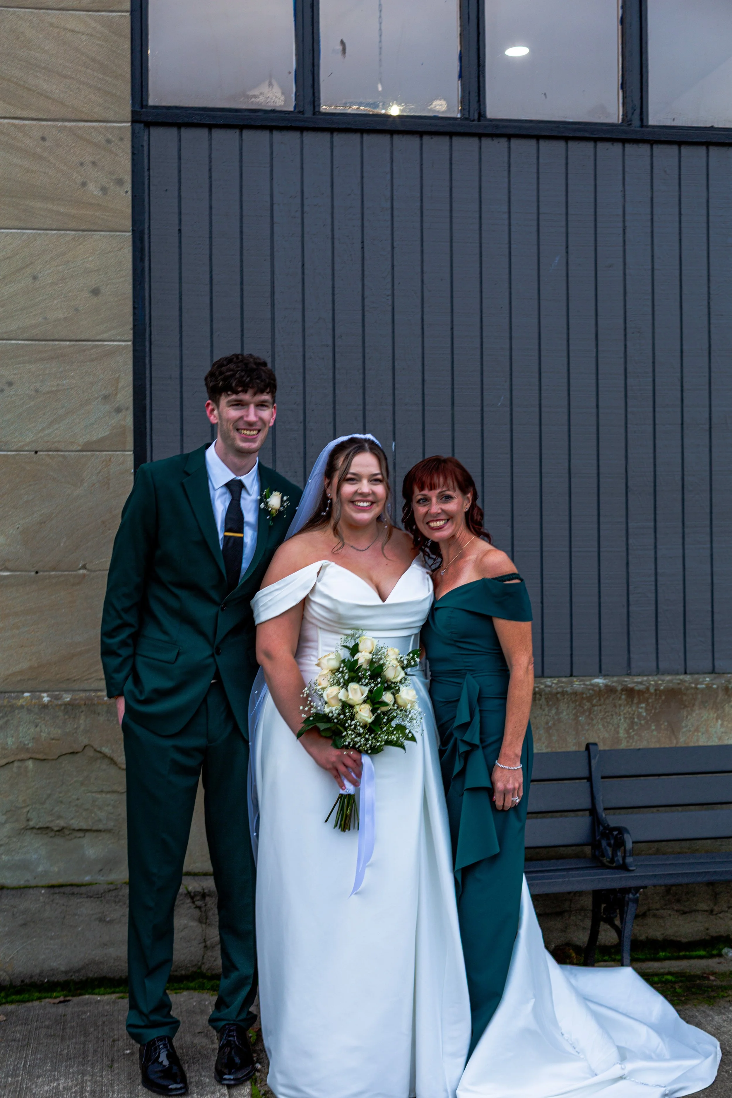 A bride in a white wedding dress holding a bouquet, standing between a groom in a dark suit and a woman in an teal dress, smiling in front of a gray wall and a bench.