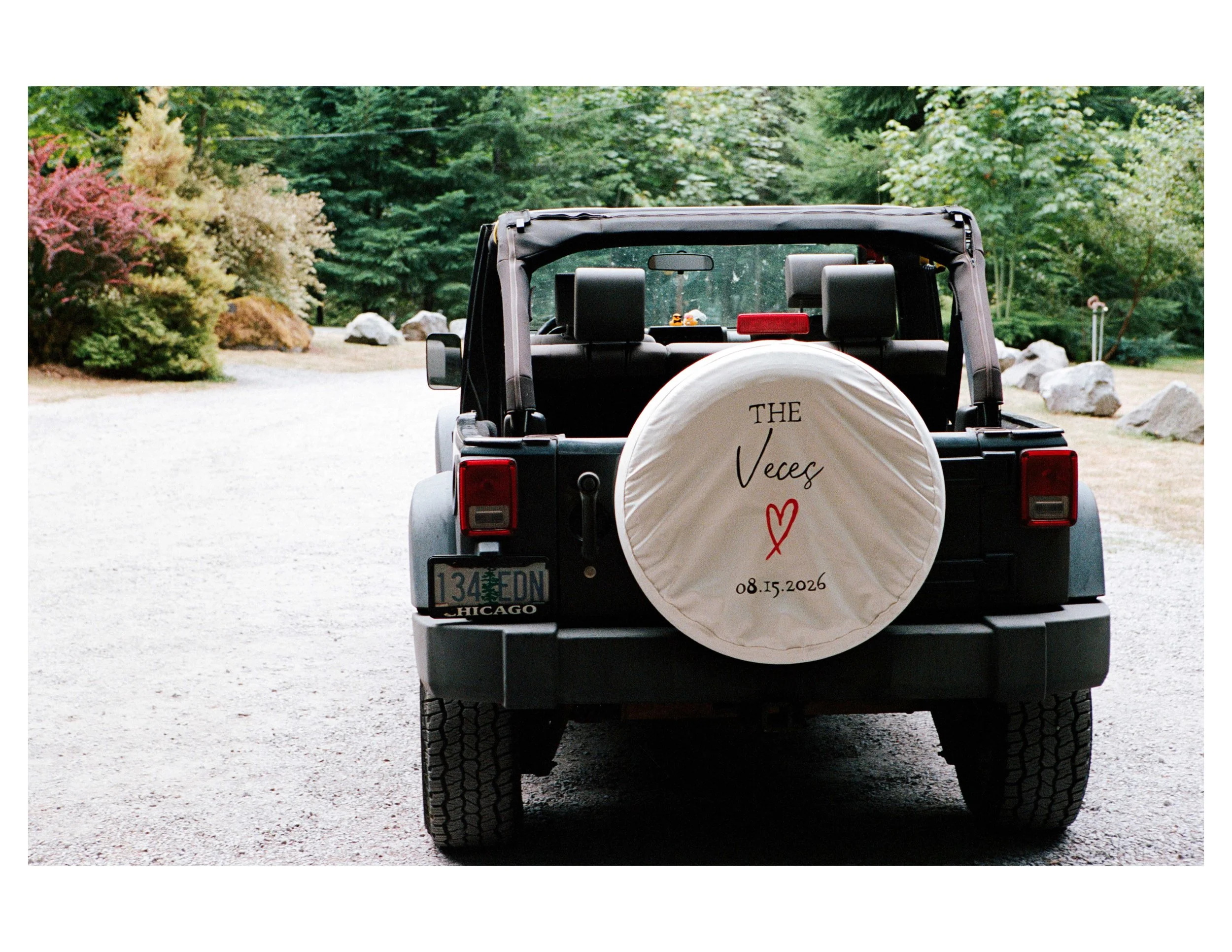 Back view of a black off-road vehicle with a spare tire cover that says "THE VeeC" and a date, parked on a gravel area with green trees in the background.