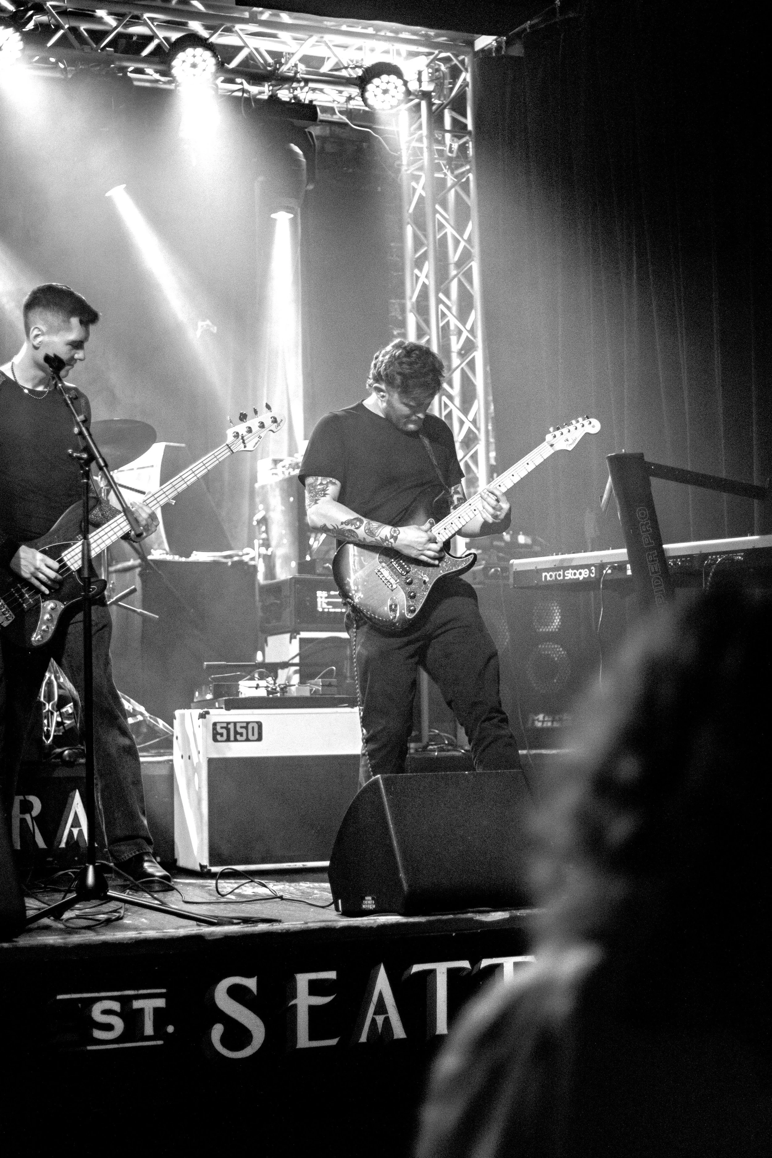 Two musicians perform on stage with electric guitars, surrounded by stage lights and equipment, during a live music concert.
