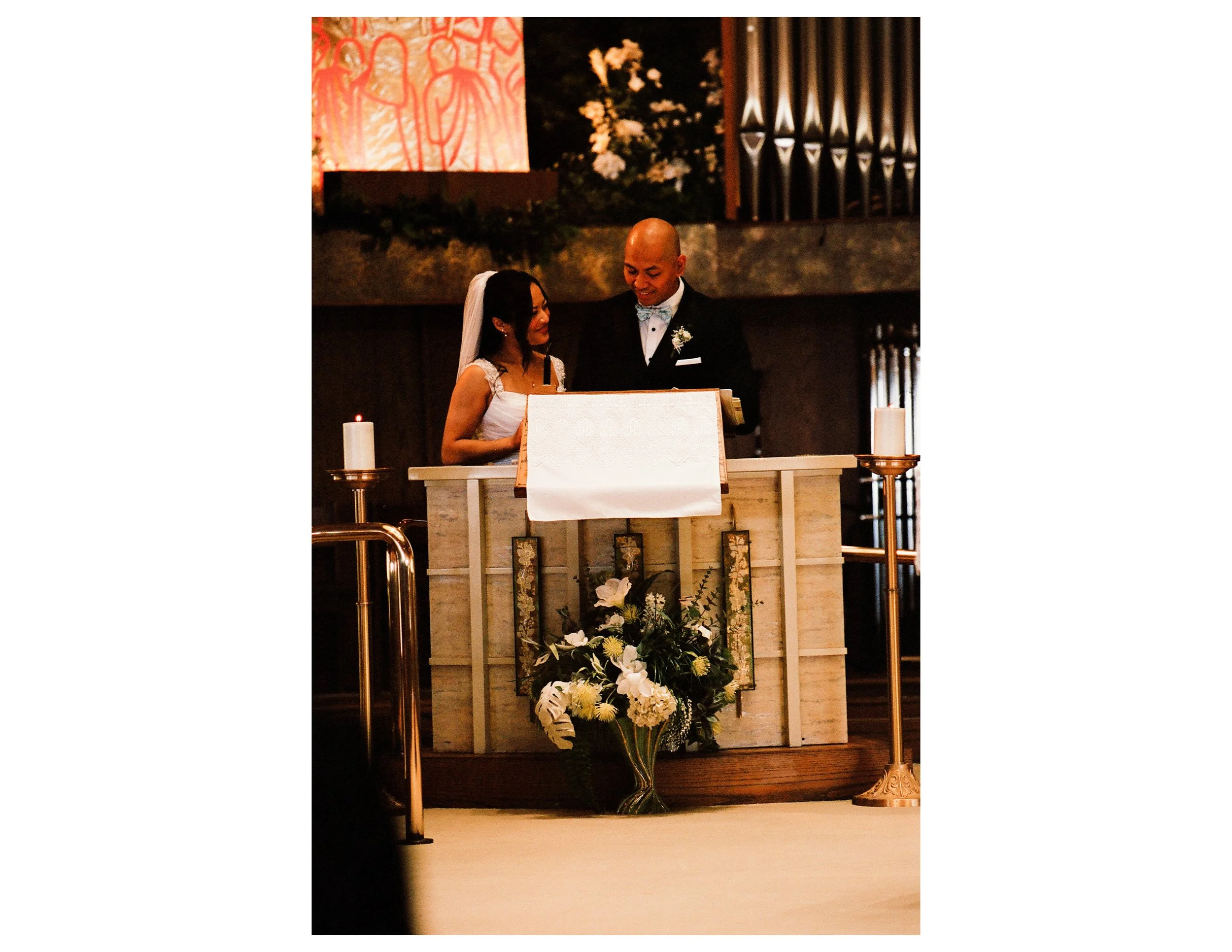 A couple standing at the altar during a wedding ceremony inside a church. The woman is wearing a white dress and veil, and the man is in a black tuxedo with a bow tie. They are smiling and looking at each other, with candles and floral arrangements a