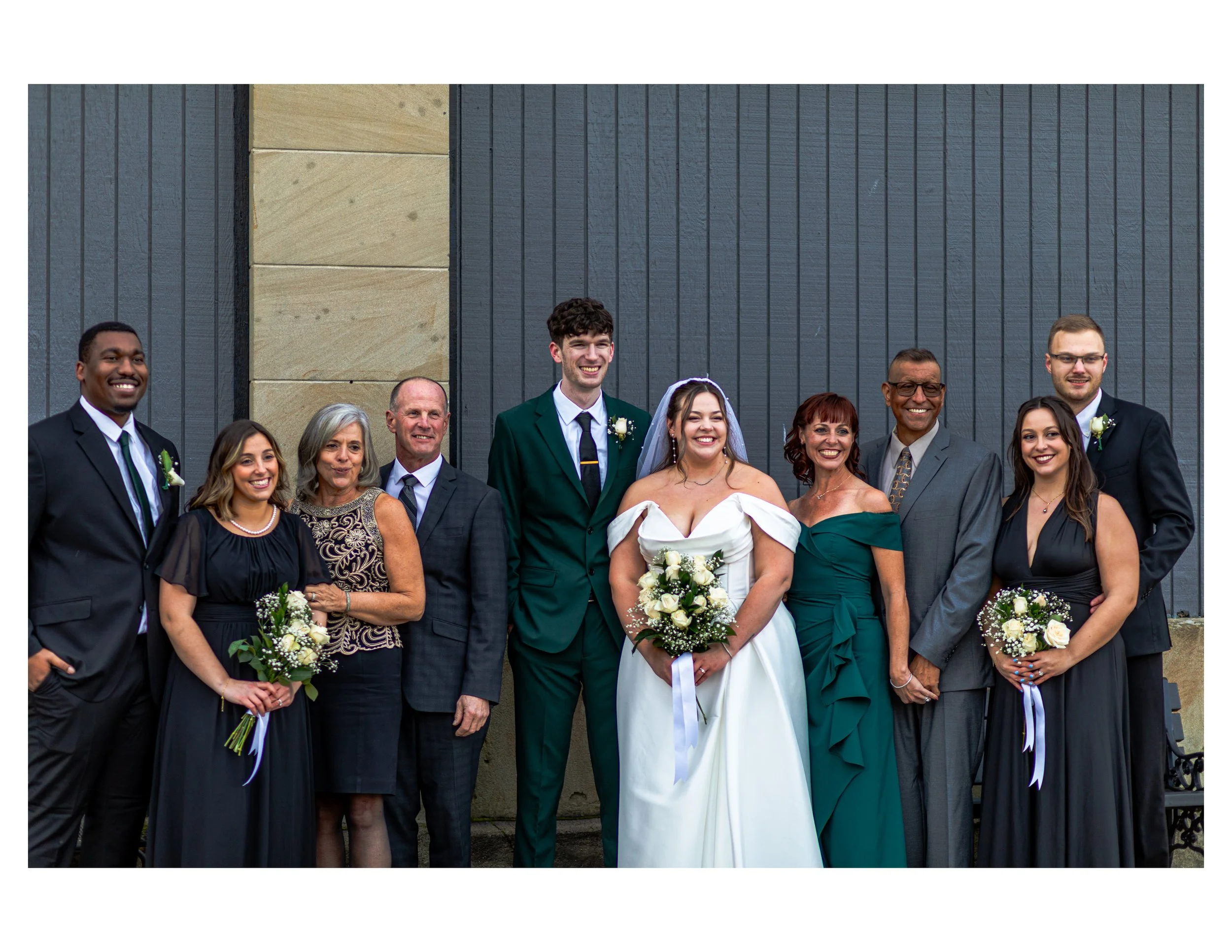 Group of diverse people dressed in formal attire, smiling, standing in front of a gray wall, likely at a wedding or celebration.