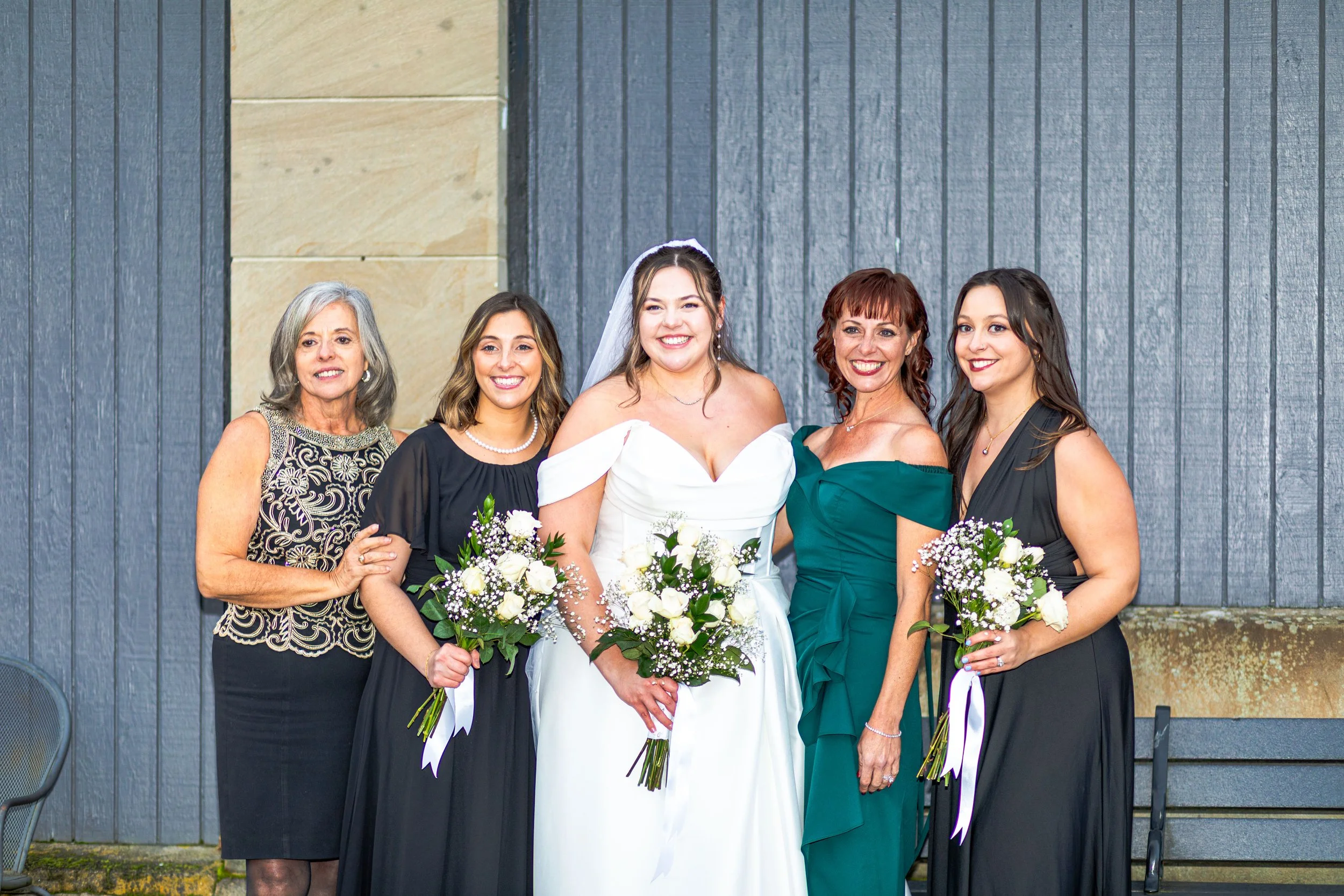 A group of five women dressed in formal attire, including a bride in a white wedding gown with a veil, standing outdoors against a blue and beige wall, smiling and holding bouquets of white flowers.