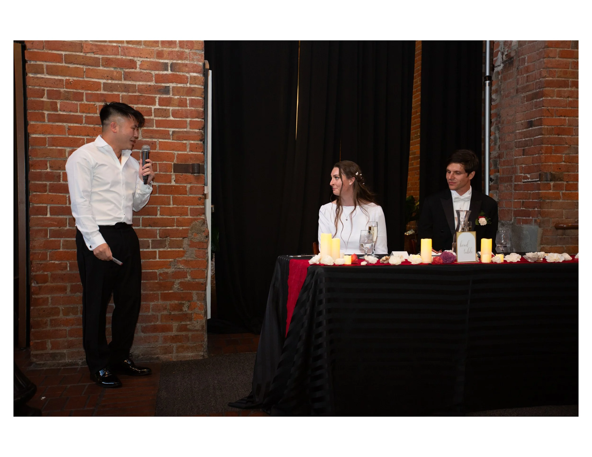 Speech at a wedding reception. Two men sitting at a table with candles and floral decor, one man standing with a microphone, both dressed in formal attire. Brick wall background.