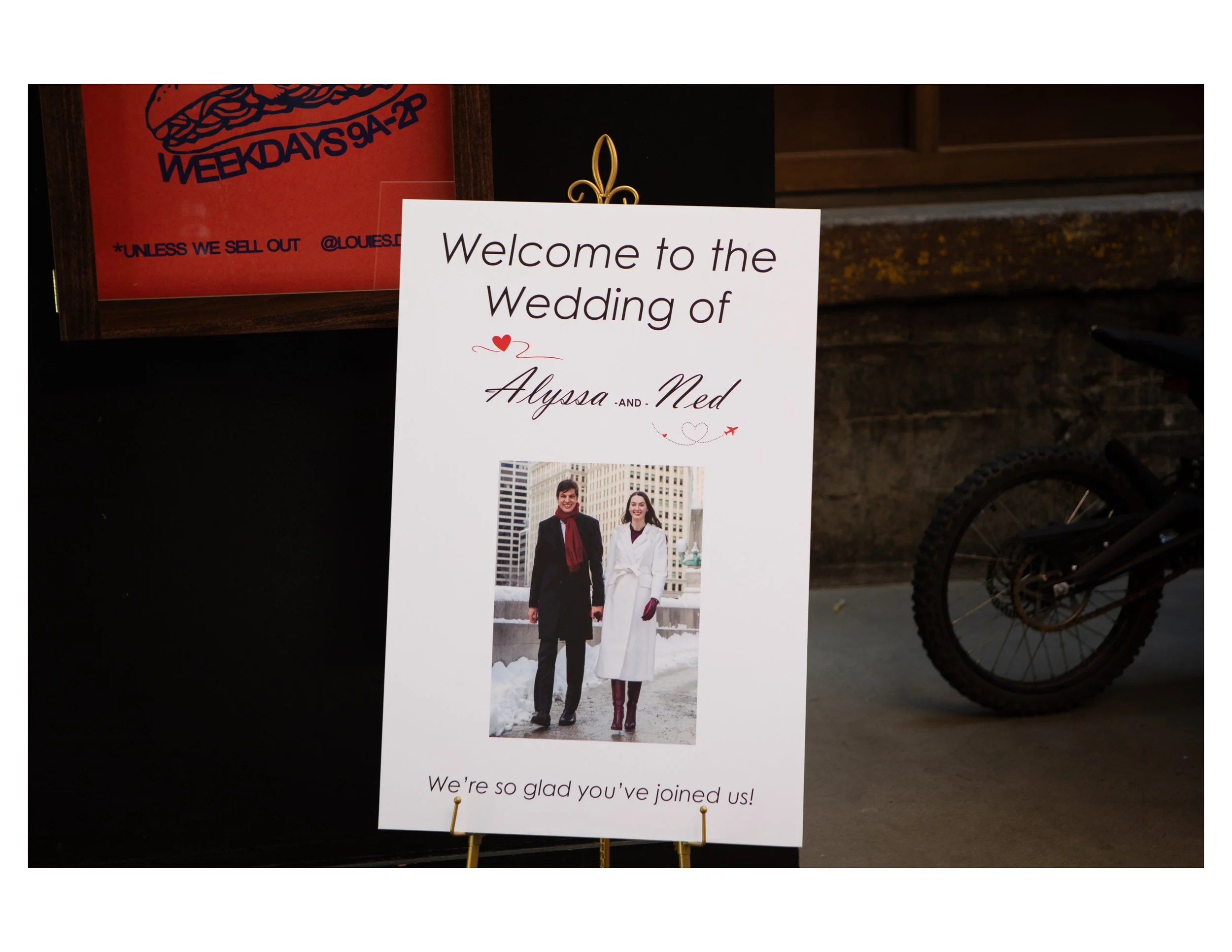 Wedding welcome sign with photo of a smiling couple in winter clothing, on a black background with a partial restaurant or cafe menu board nearby.