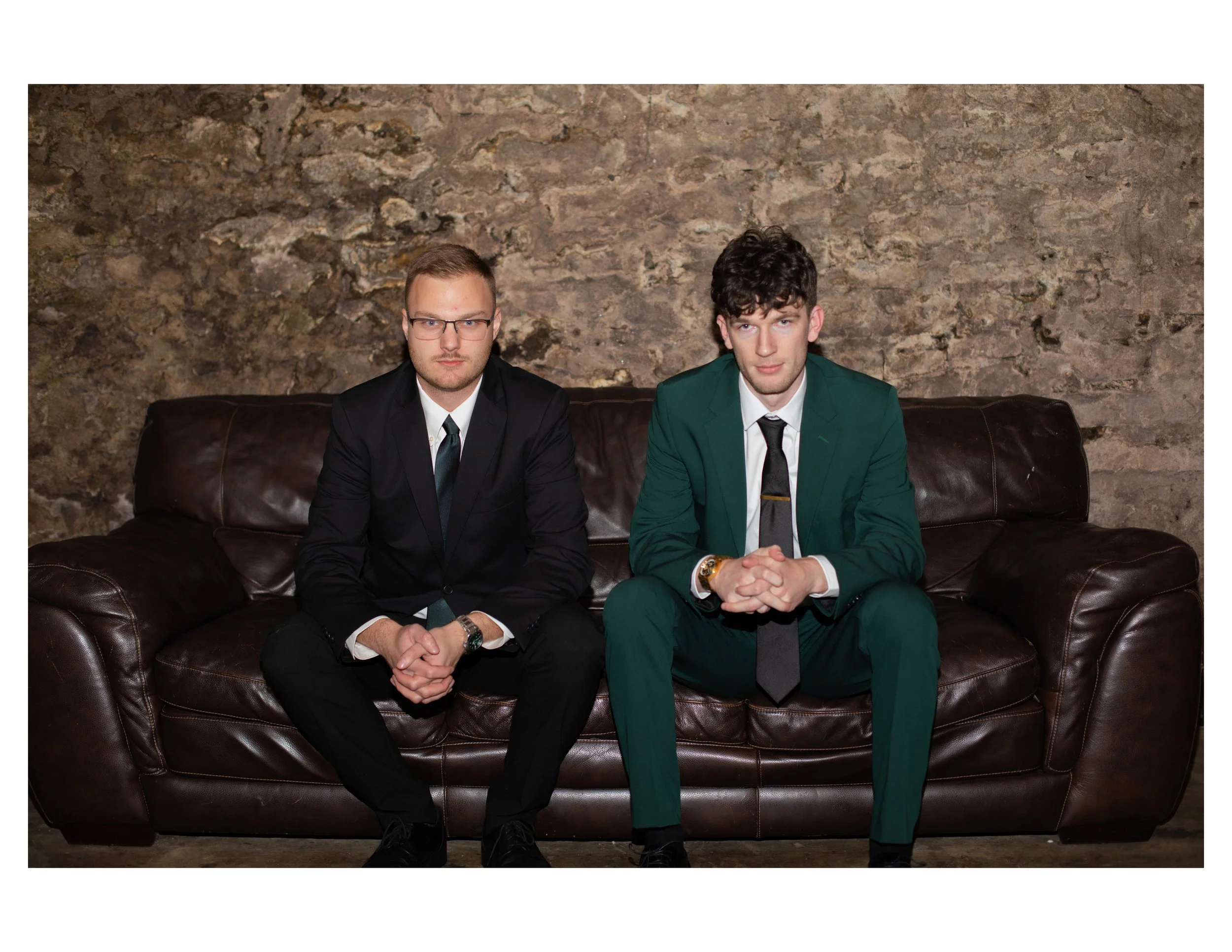 Two men in suits sitting on a leather couch in front of a stone wall.