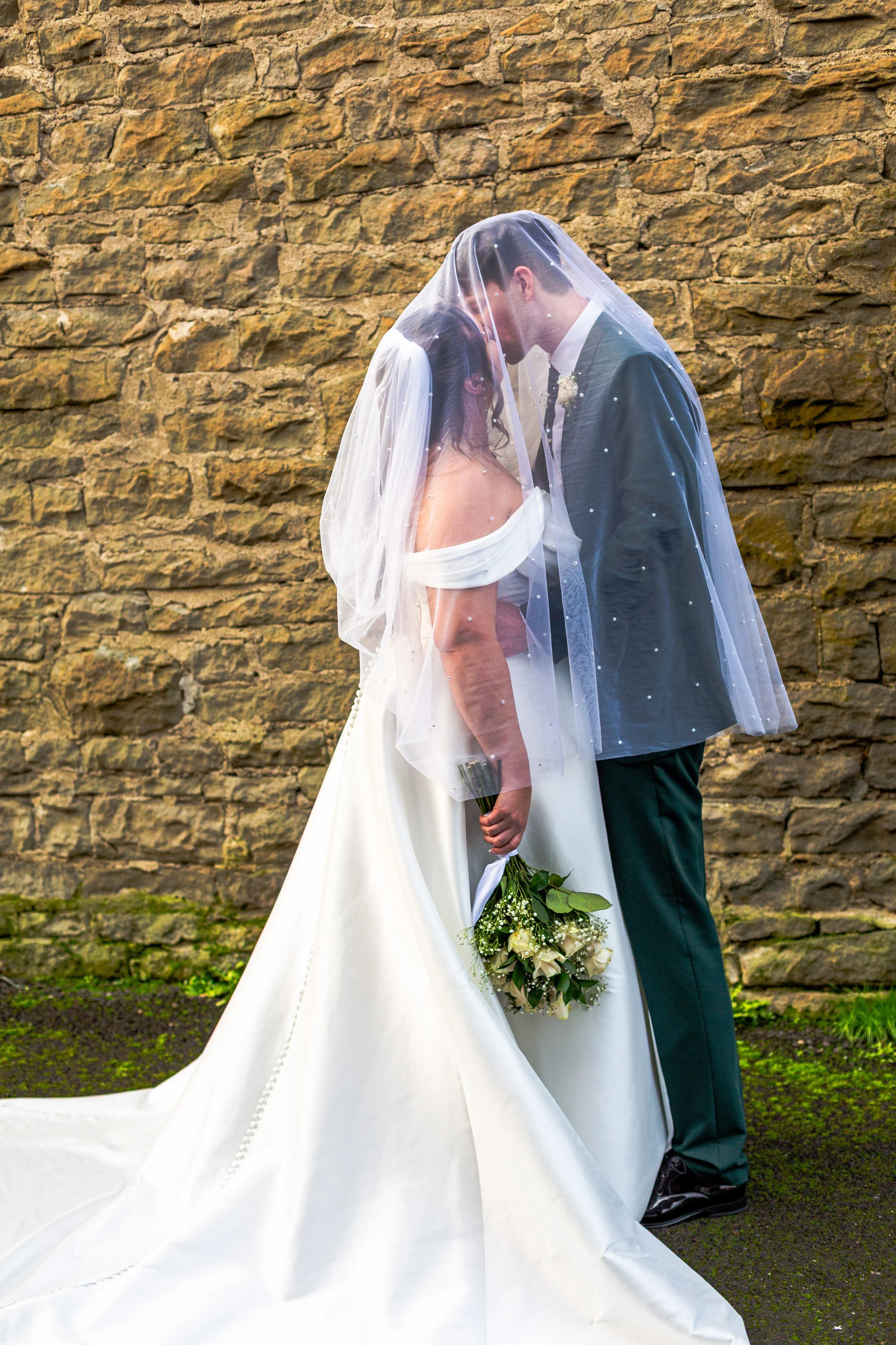 A couple dressed in wedding attire, with the bride in a white gown and the groom in a dark suit, stand close together under a sheer veil outdoors against a stone wall. The bride holds a bouquet of white roses and greenery.