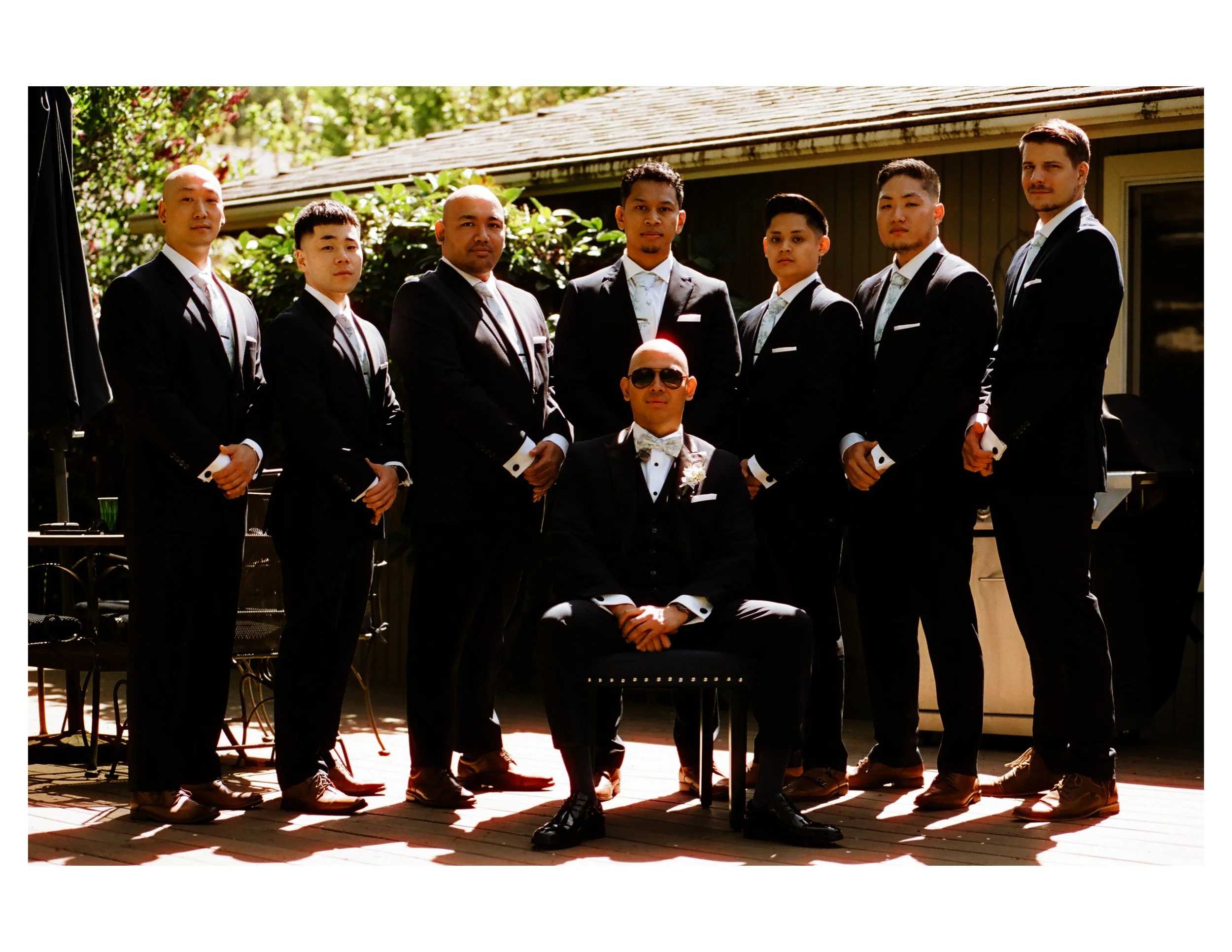 A group of eight men dressed in formal tuxedos, standing outdoors in front of a house with greenery, during daytime, with one seated man wearing sunglasses in the foreground.