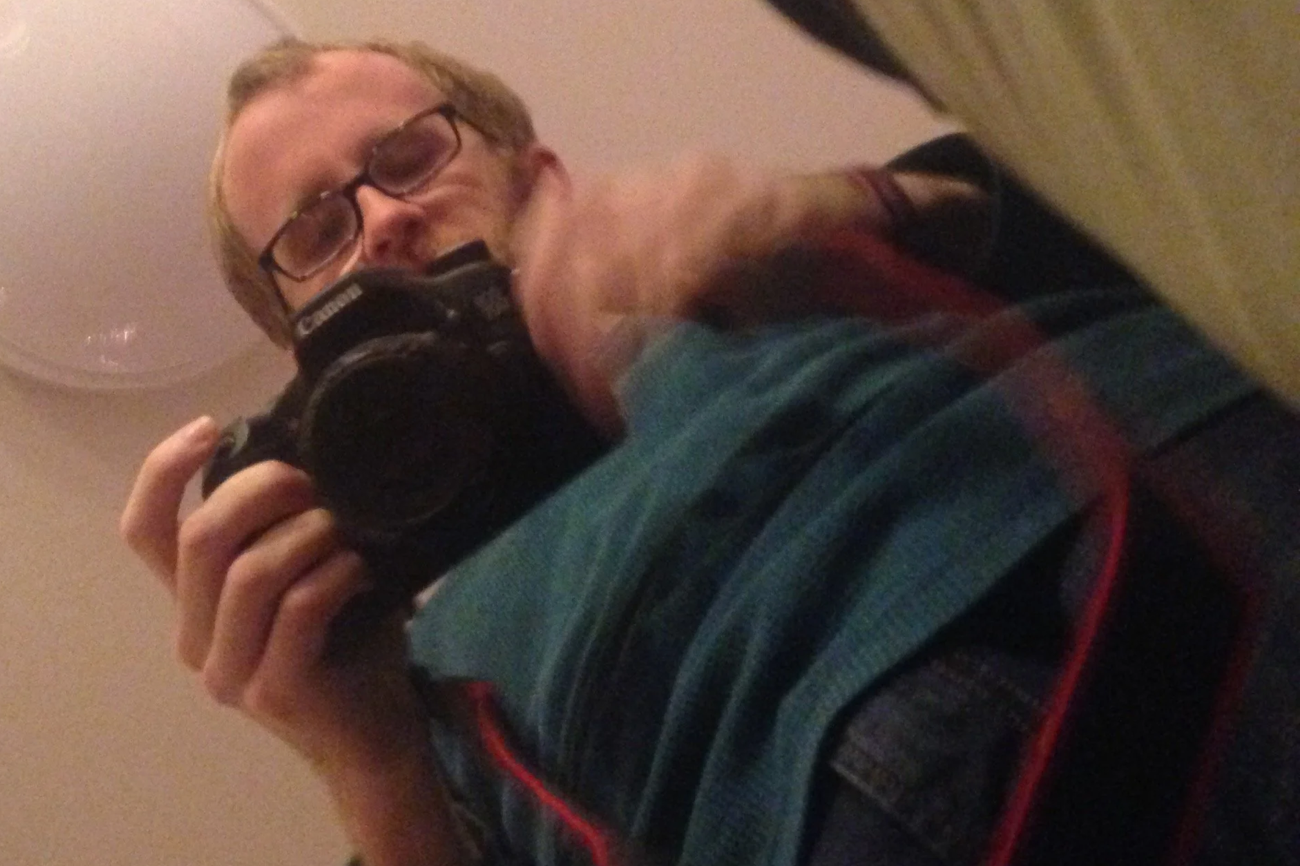 Person taking a selfie with a camera, wearing glasses and a striped hoodie, with a ceiling light visible in the background.