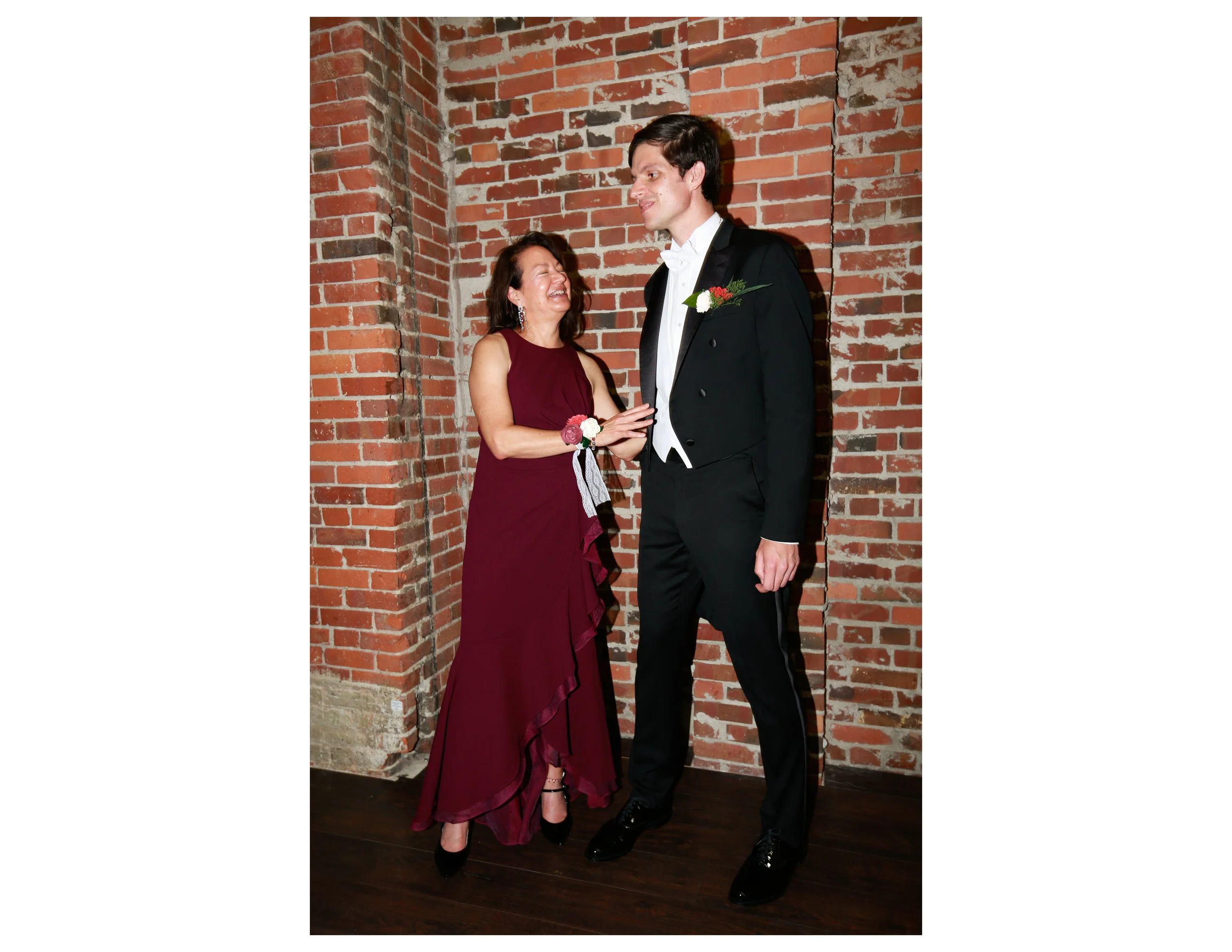A woman in a burgundy dress and a man in a black tuxedo with a white bow tie standing in front of a brick wall, smiling and holding hands.