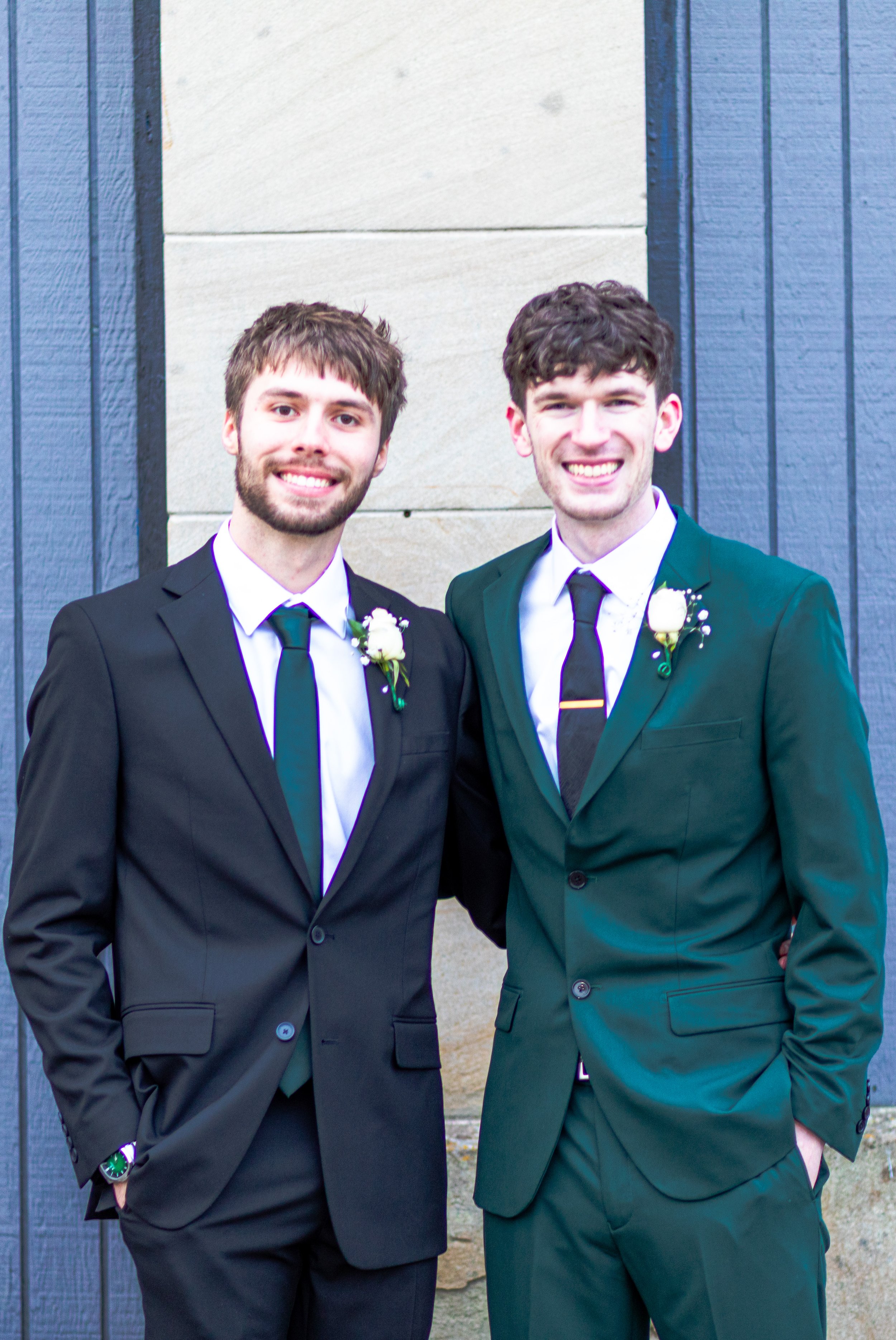 Two young men in formal suits, each with a white boutonniere and tie, smiling and posing outdoors in front of a blue and beige wall.