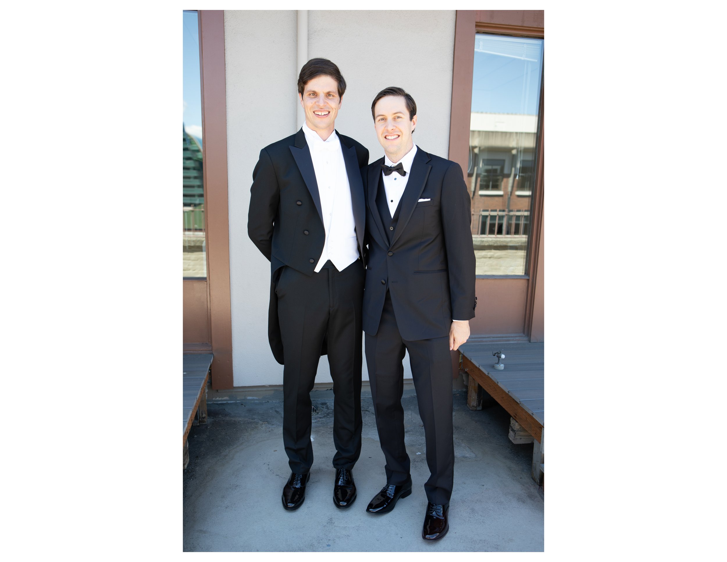 Two men dressed in black tuxedos and white dress shirts standing side by side outdoors in front of a building with large windows, smiling at the camera.
