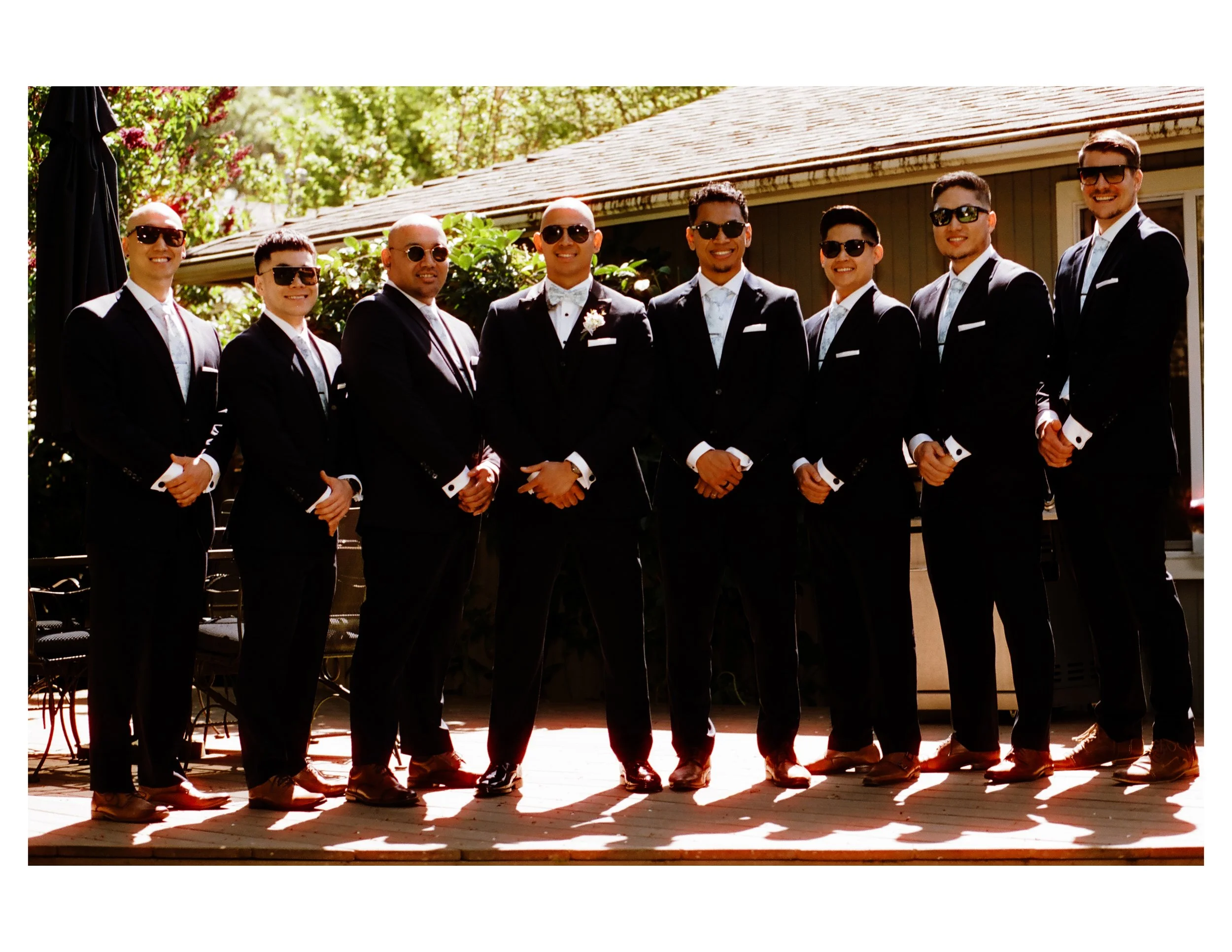 A group of ten men dressed in black tuxedos with white shirts and black sunglasses, standing outdoors on a wooden deck during daytime, smiling for a photo.