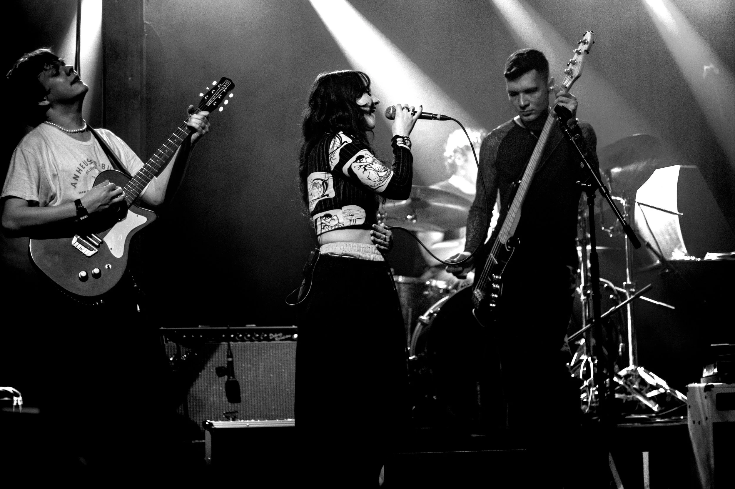 A black-and-white photo of three musicians performing on stage, with a guitarist on the left, a singer in the middle holding a microphone, and a bassist on the right. A drum set and music stand are visible in the background.