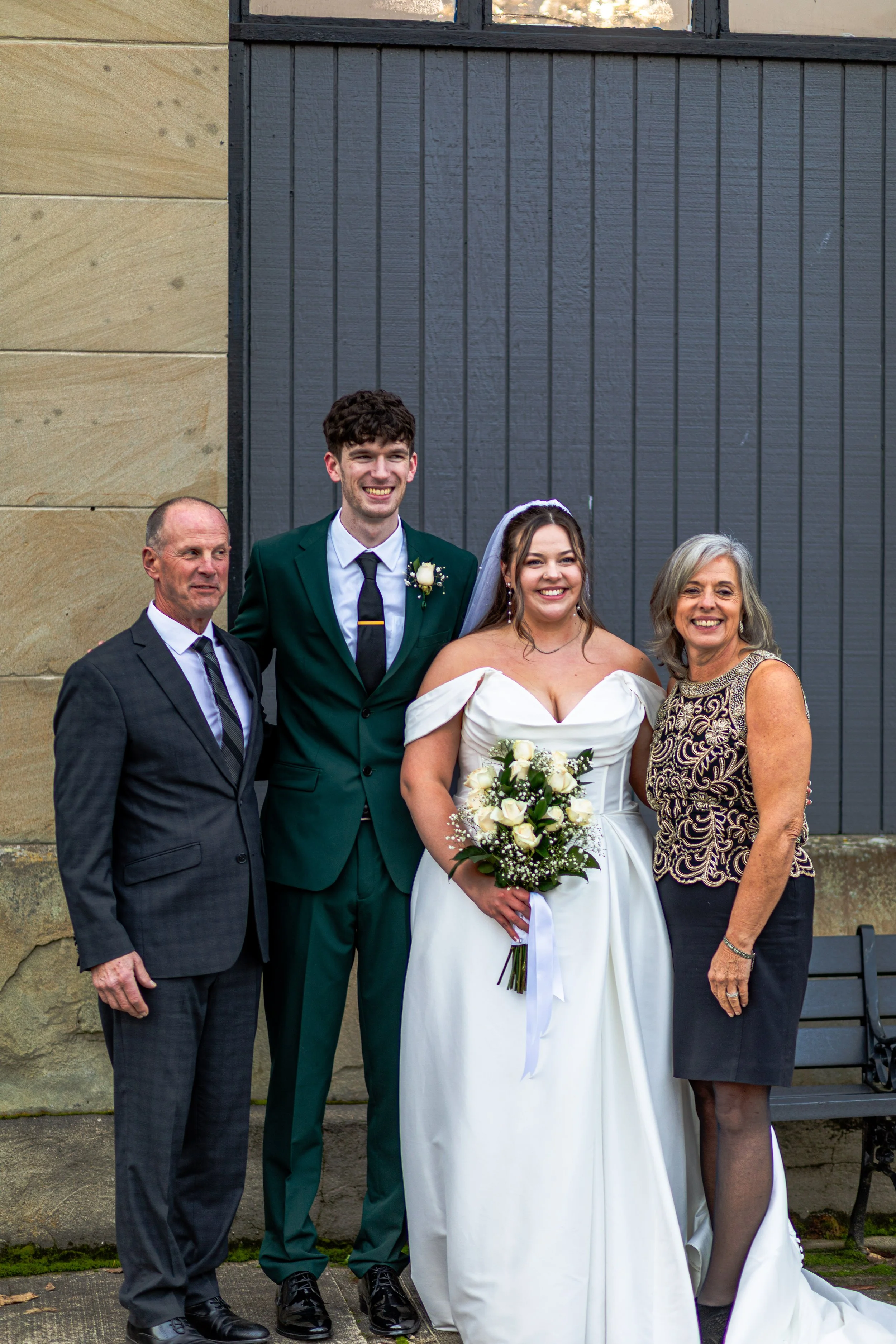A group of four people at a wedding, including a bride in a white dress holding a bouquet of white roses, a groom in a green suit, and two older adults, all smiling and standing outdoors in front of a building with a stone wall and dark grey wooden p