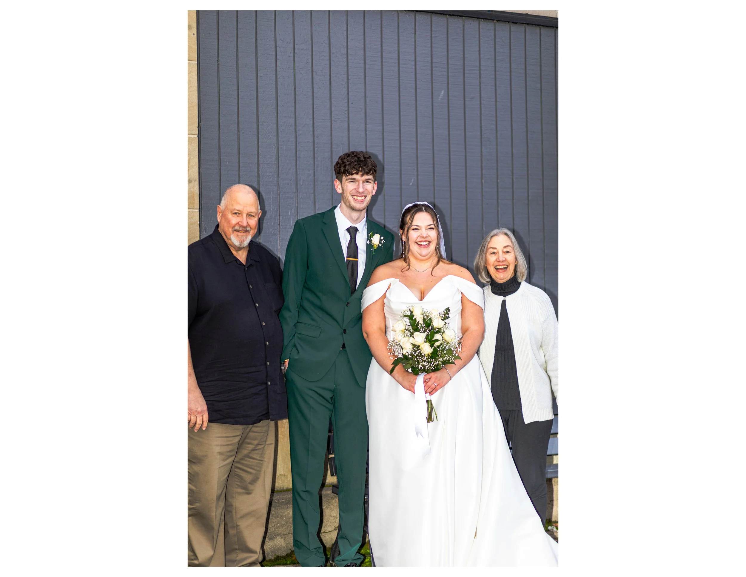 A group of four people standing outdoors, celebrating a wedding. The bride is in a white dress holding a bouquet, and the groom is in a green suit with a white boutonnière. Two older adults, a man and a woman, are standing on either side of the coupl