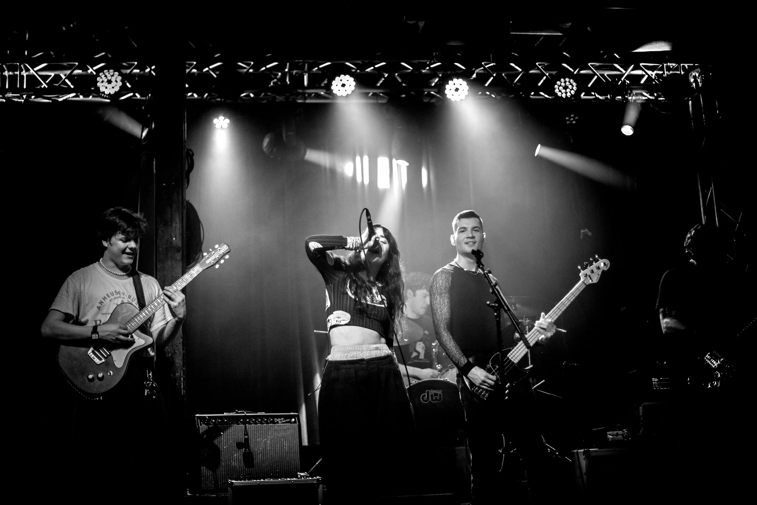 A black and white photo of a band performing on stage with a singer, guitarist, bassist, and drummer under stage lights.