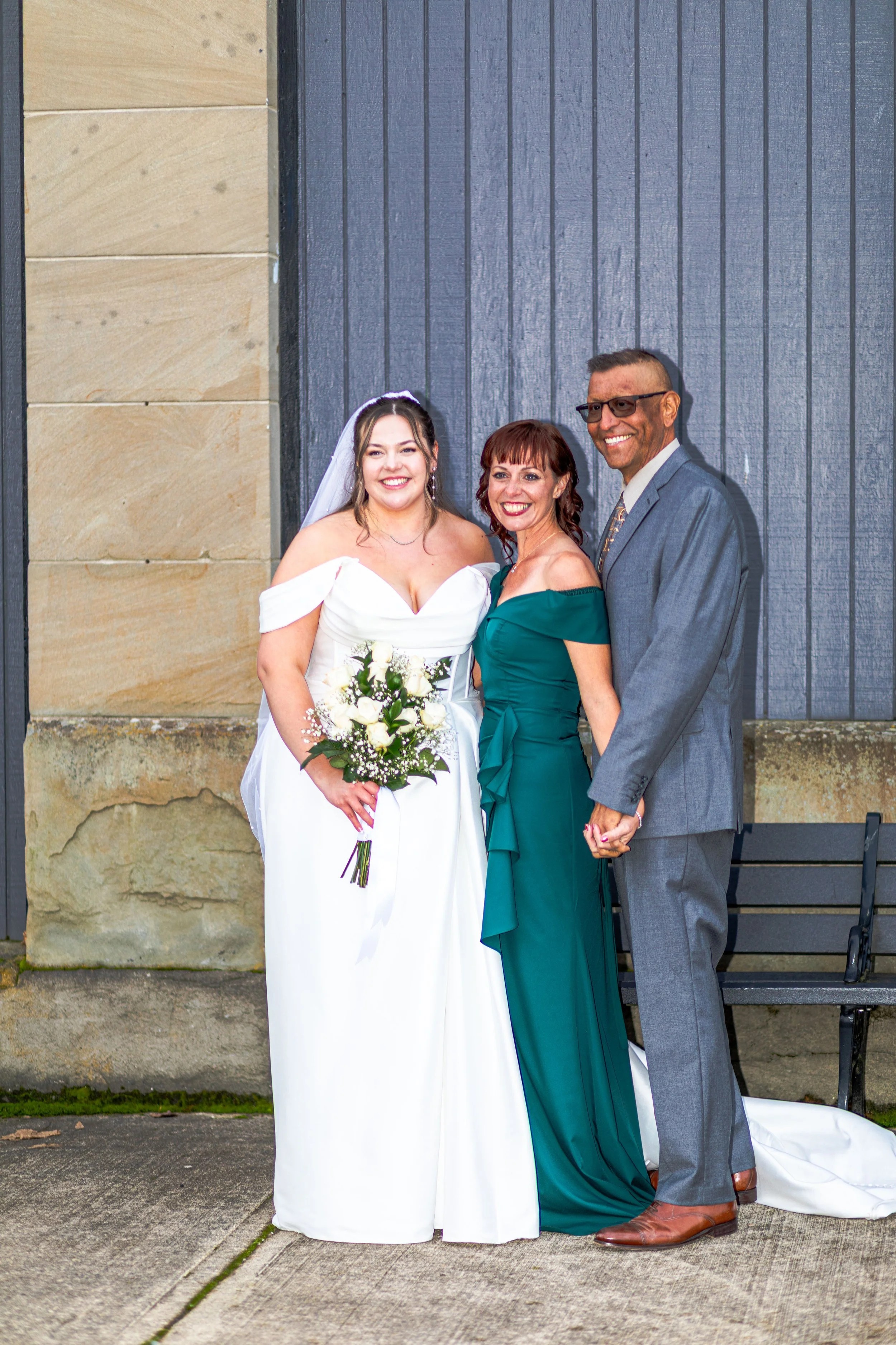 A bride in a white wedding dress holding a bouquet, standing with a woman in a teal dress and a man in a gray suit, all smiling and holding hands outdoors near a building with a stone and wood wall.