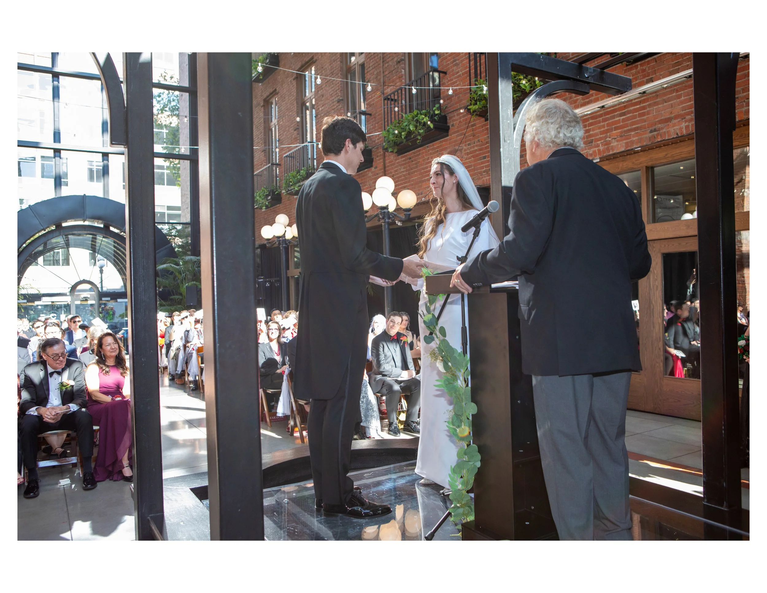 A wedding ceremony taking place outdoors in an urban setting, with a groom and bride exchanging vows while an officiant conducts the ceremony. Guests watch, seated and standing, in front of a brick building decorated with string lights and greenery.