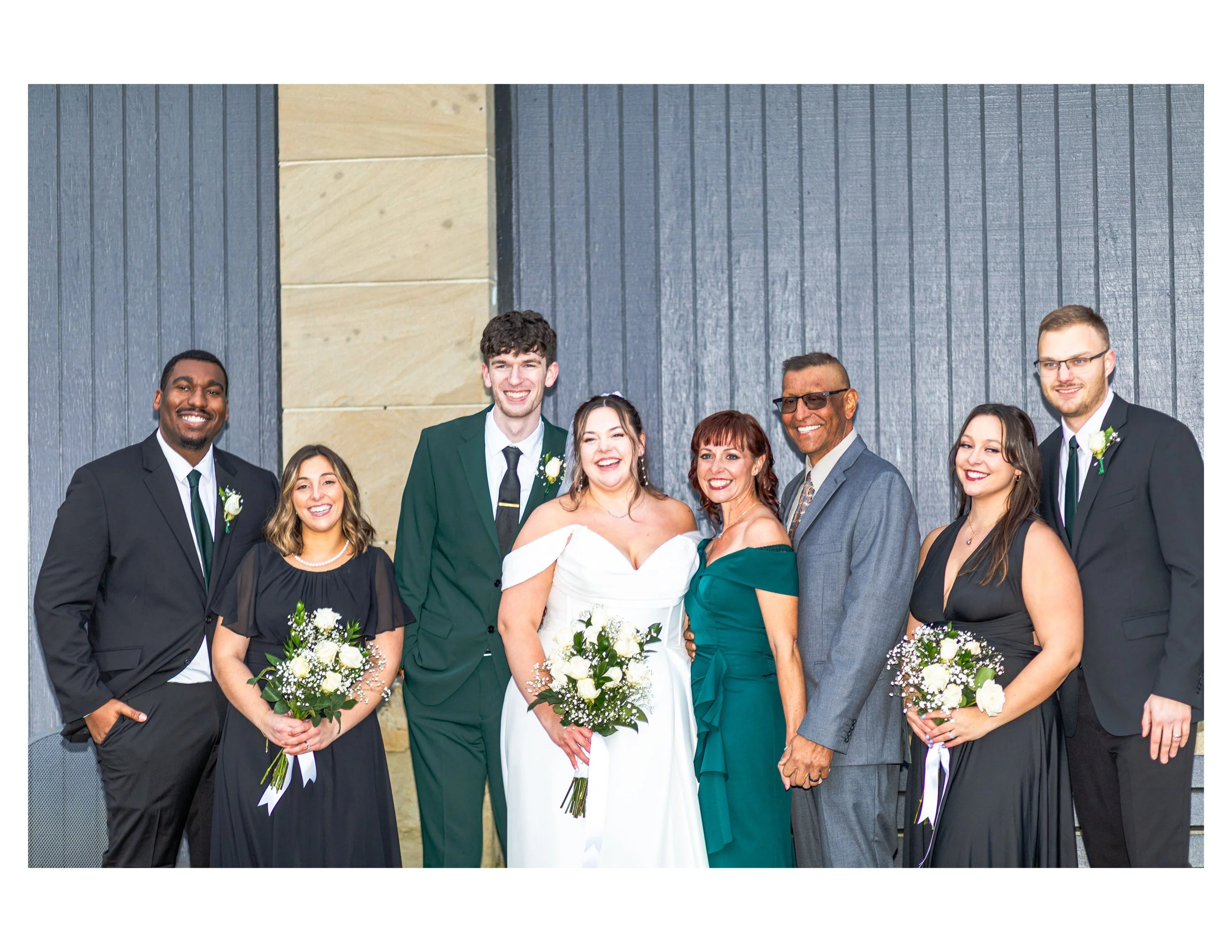 A group of people dressed in formal attire, posing for a wedding photo outdoors in front of a gray and beige wall.