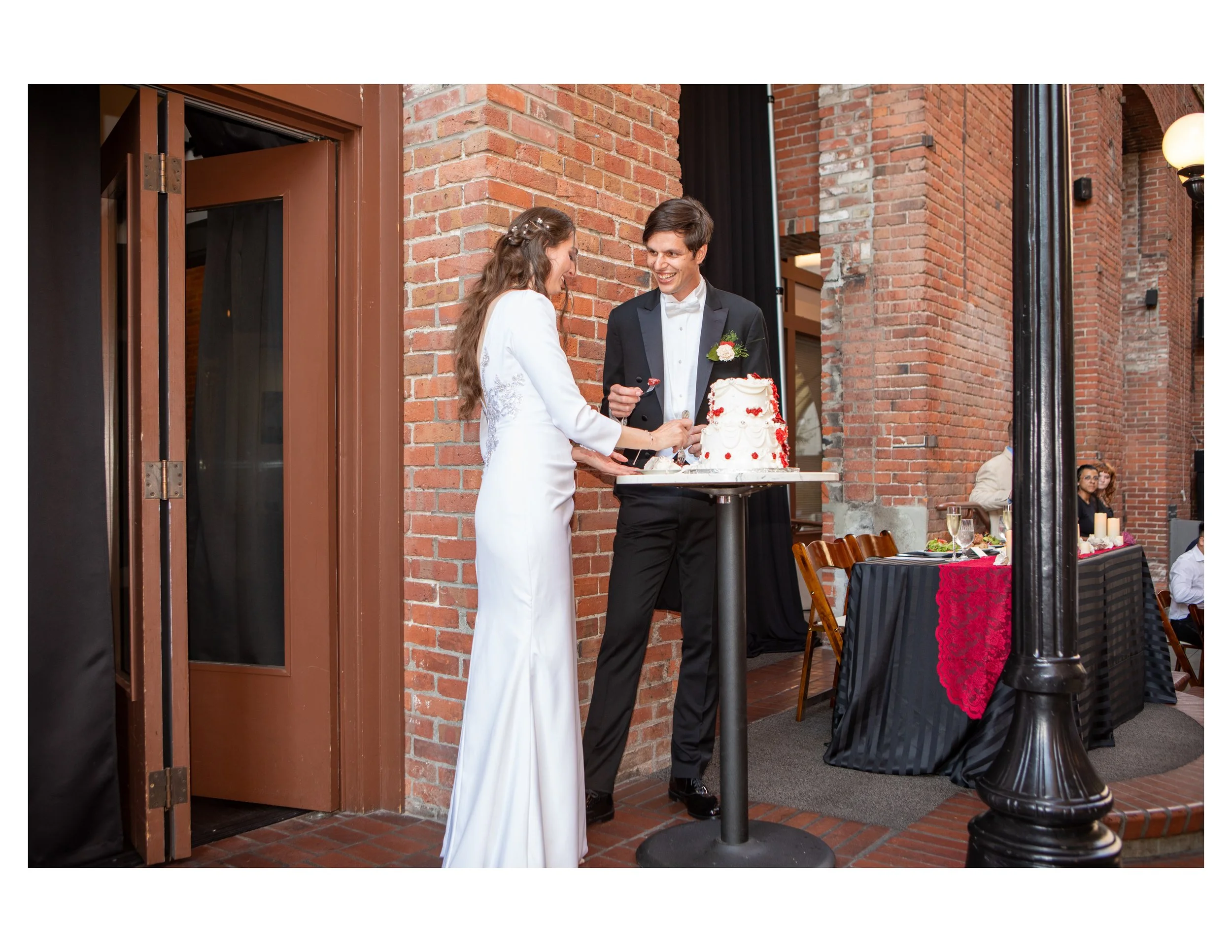 A bride and groom standing in front of a brick wall, smiling and holding a cake. The bride wears a white dress, and the groom wears a black tuxedo with a white bow tie and boutonniere. The event appears to be a wedding reception.