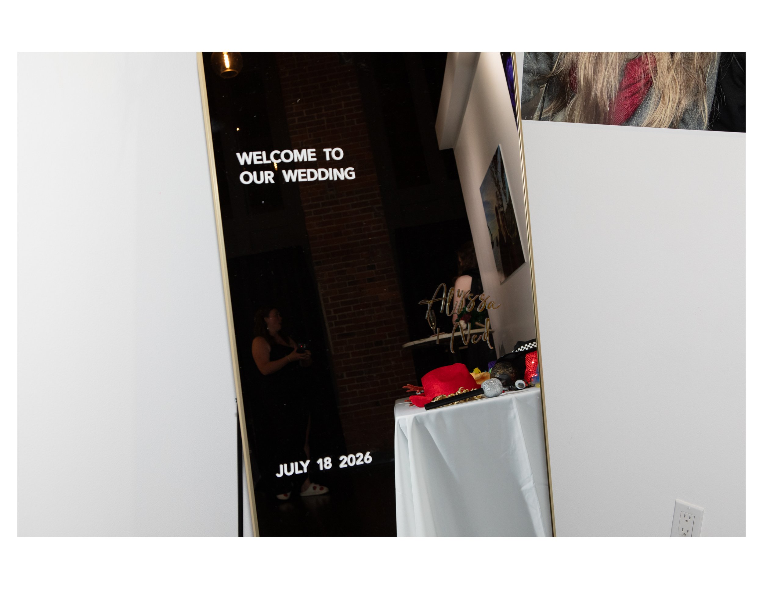 A wedding welcome sign in black with white text, reflecting in a mirror, with a date of July 18, 2026, next to a table decorated with red and gold items.