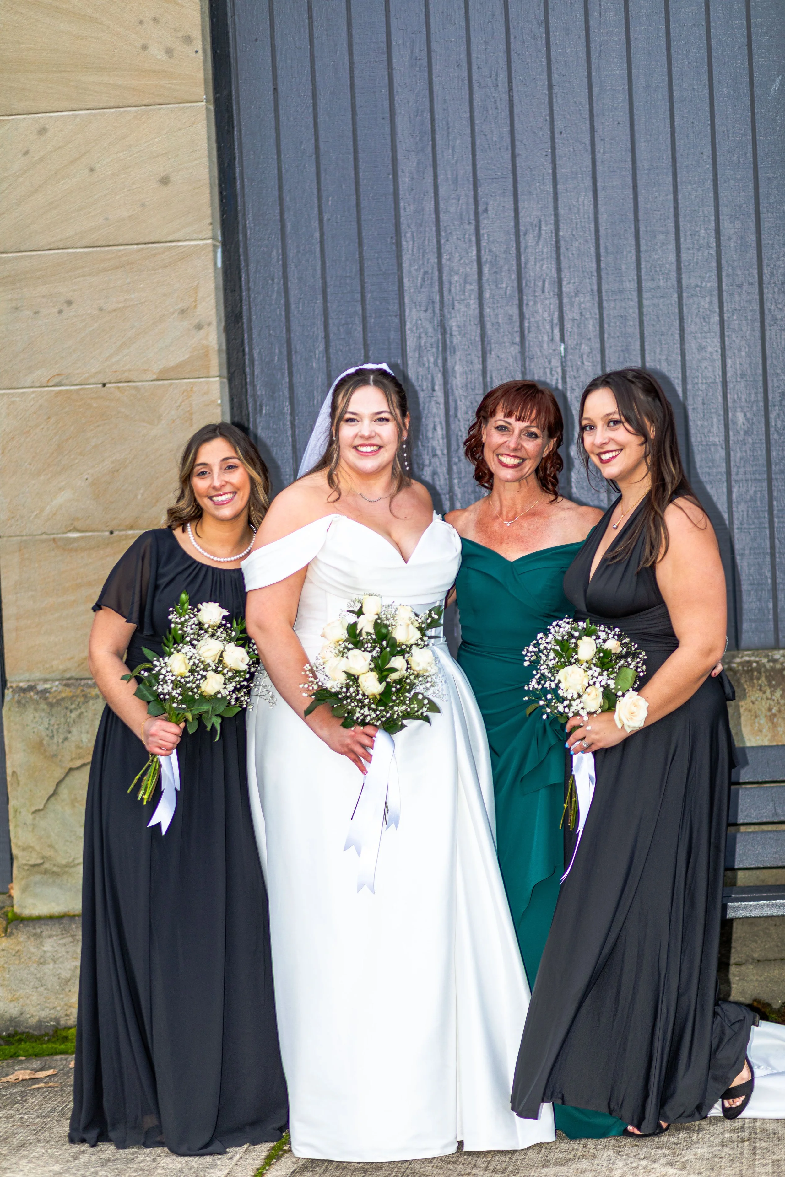 A bride in a white wedding dress with off-the-shoulder sleeves stands between three women, all holding bouquets of white roses and greenery. The women wear black and green dresses. They are standing outside against a textured wall with a large door.