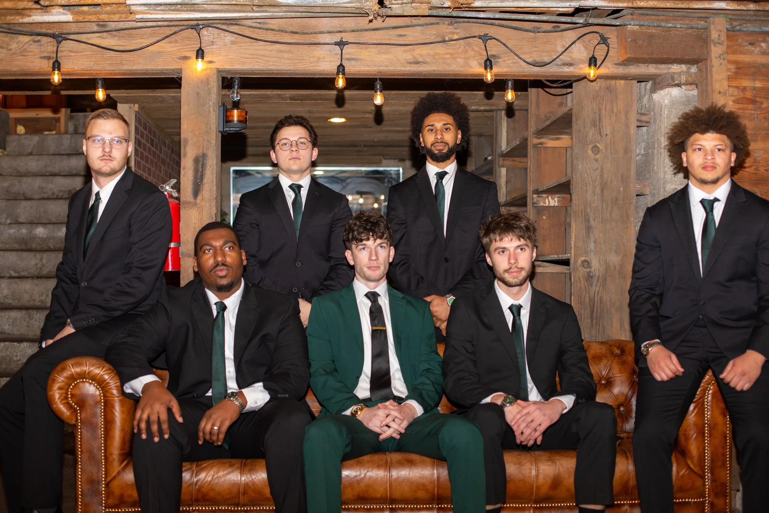 A group of nine men dressed in formal suits, some with ties, posing in a rustic indoor setting with wooden beams and string lights.