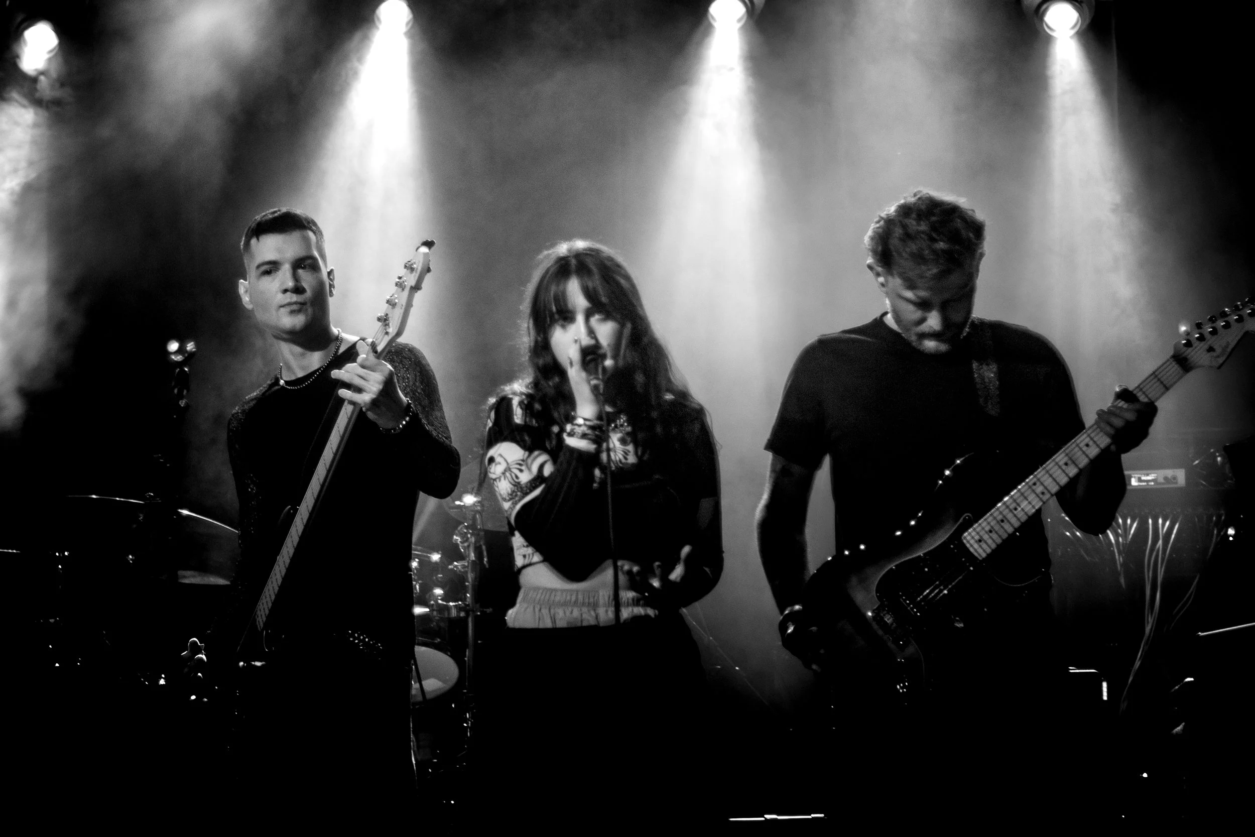 Black and white photo of a band performing on stage, with a male guitarist on the left, a female vocalist in the center, and another male guitarist on the right, with stage lights overhead.