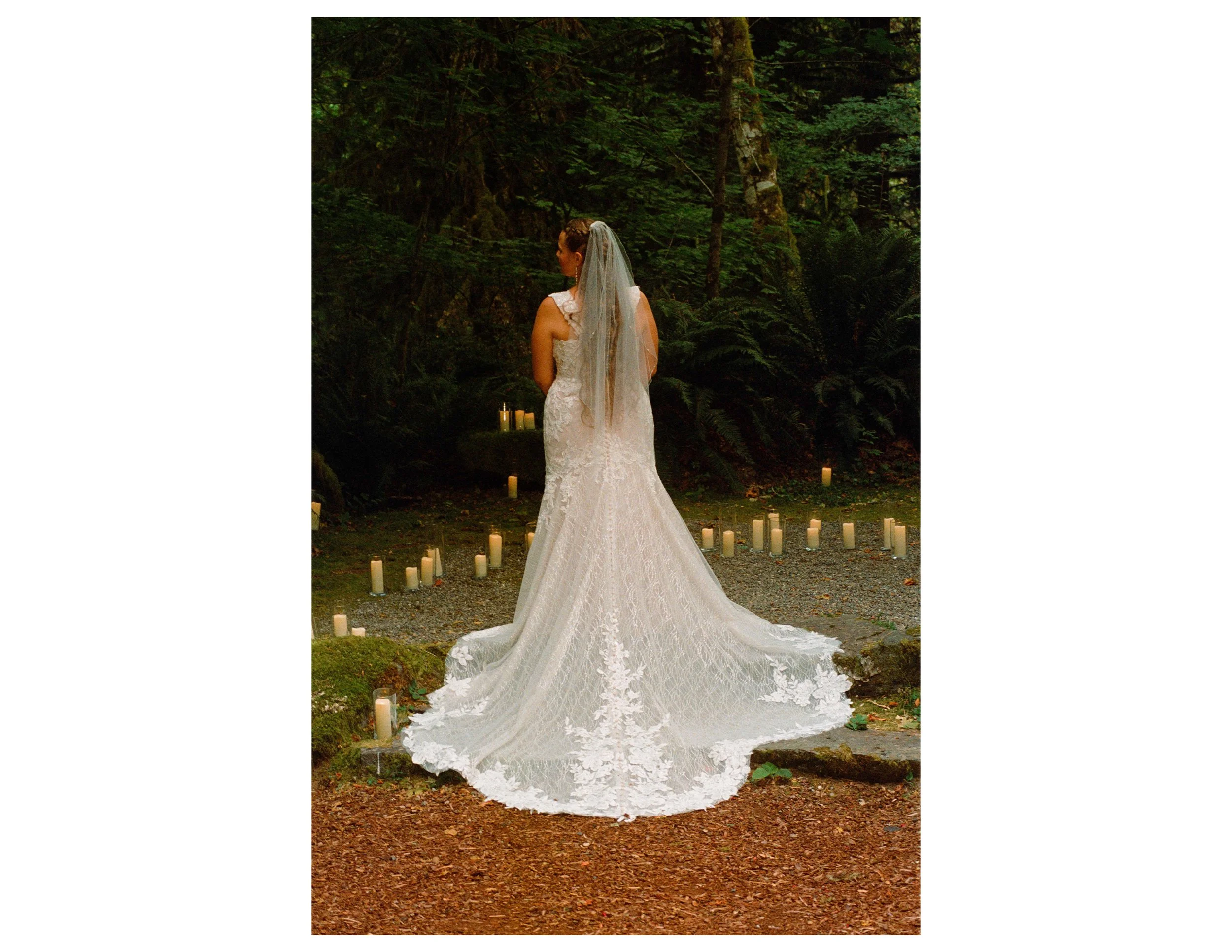 A woman in a white wedding dress with a veil, standing outdoors in a forested area surrounded by candles on the ground.