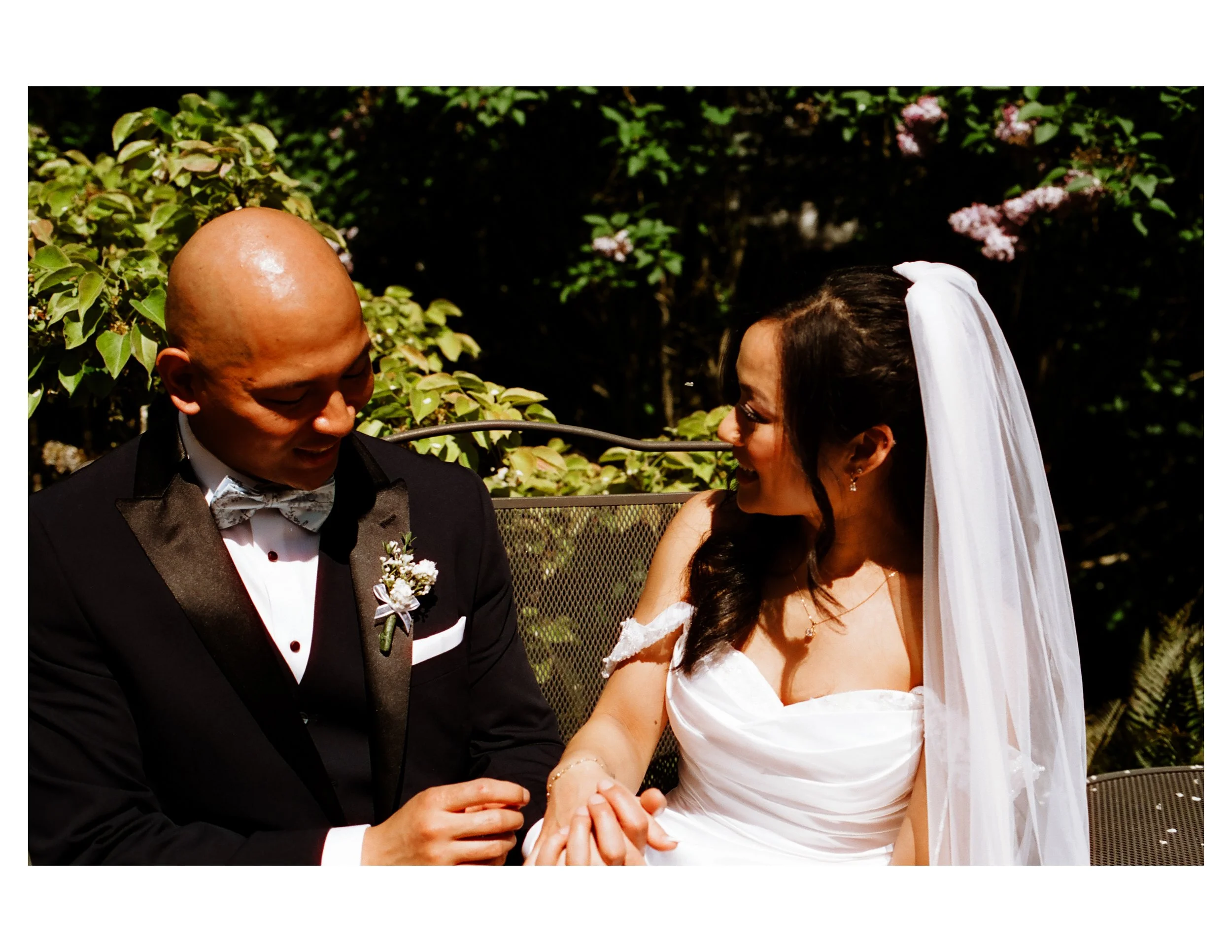 A bride and groom holding hands during their wedding ceremony outdoors with greenery and flowering bushes in the background.