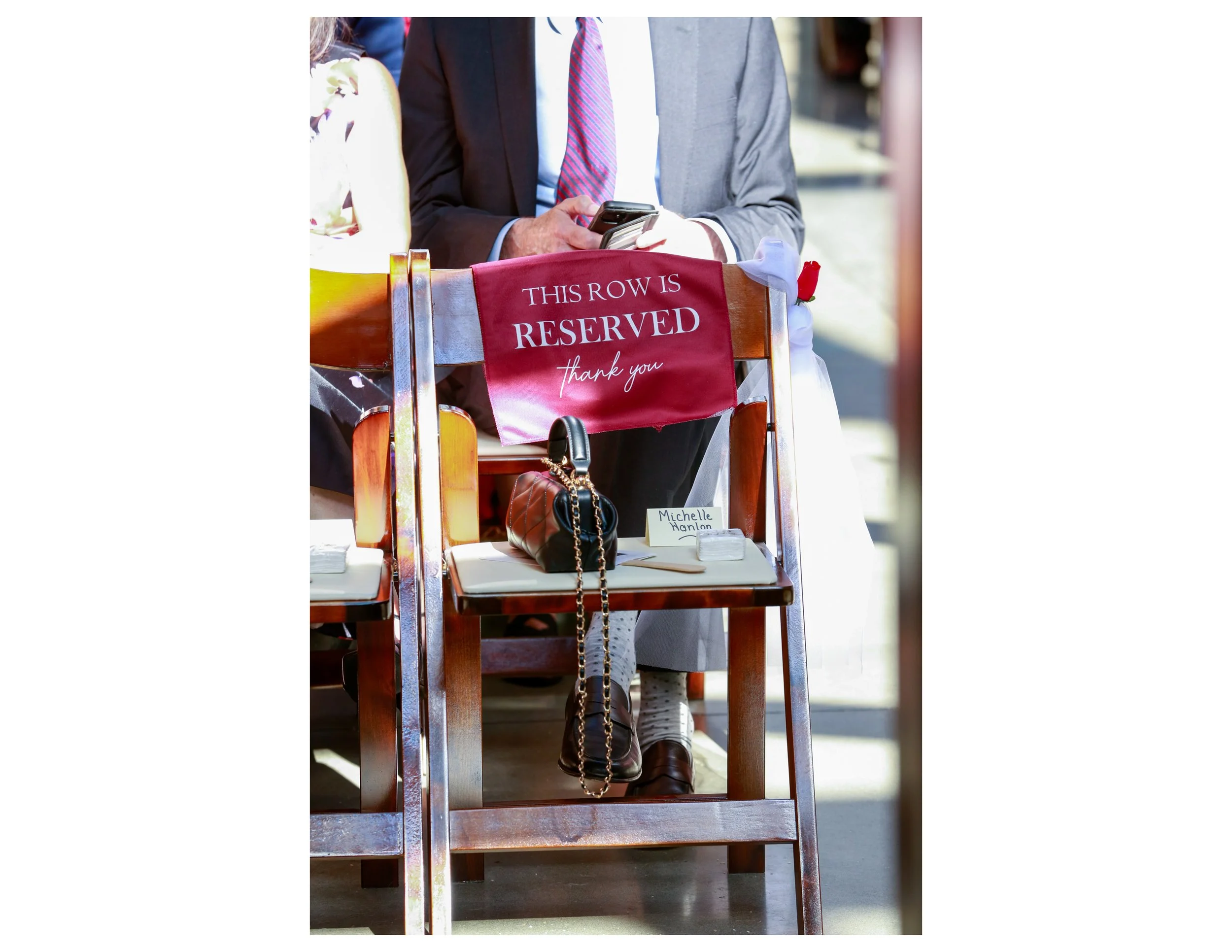 A close-up of a person's hands holding a smartphone, sitting on a reserved row at an event. The reserved sign is red with white lettering, reading 'This row is reserved, thank you.' The person is dressed in a suit with a tie, and there's a handbag an