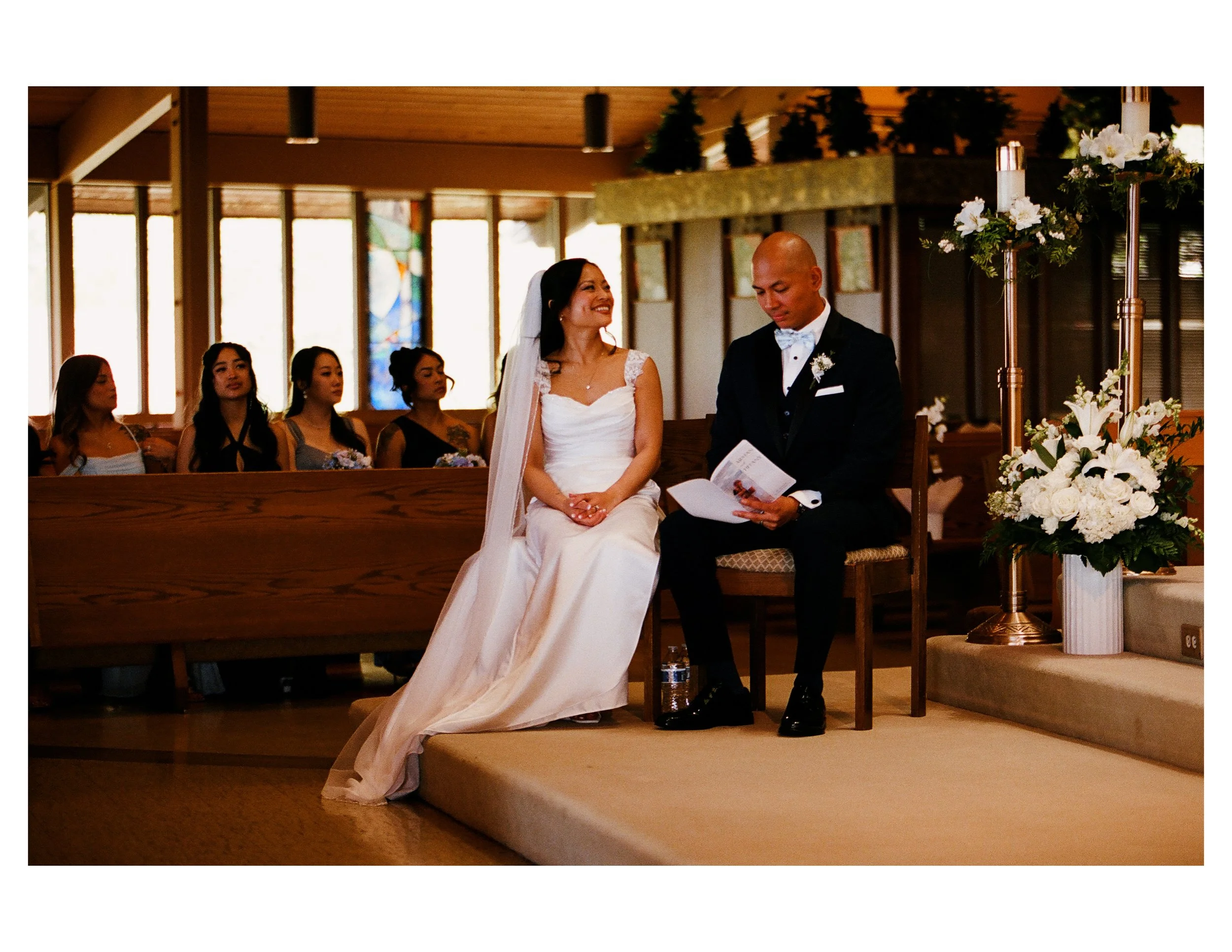 A bride and groom seated during a wedding ceremony in a church, with bridesmaids visible in the background, and floral arrangements around the altar.