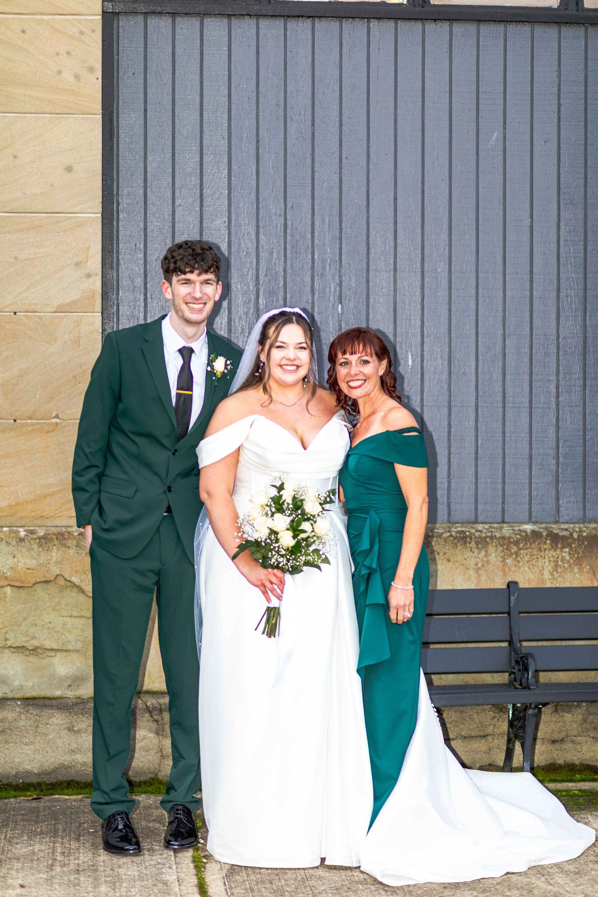 Wedding photo of three people, including a bride holding a bouquet, standing with a smiling groom in a green suit and a woman in a green dress in front of a dark wooden background.