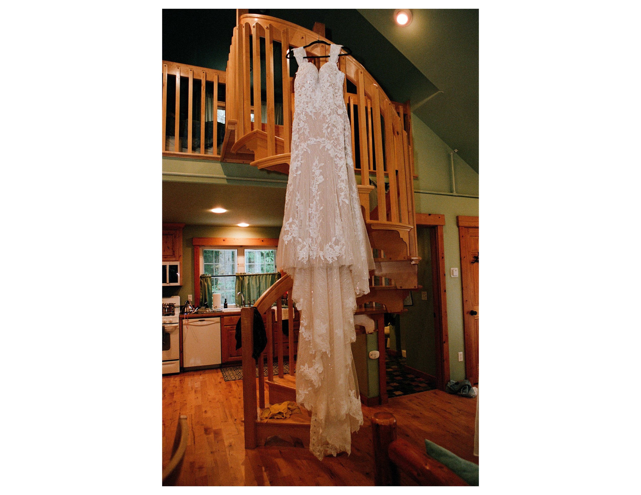 A white lace wedding dress hanging from a black hanger on a wooden staircase rail inside a house.