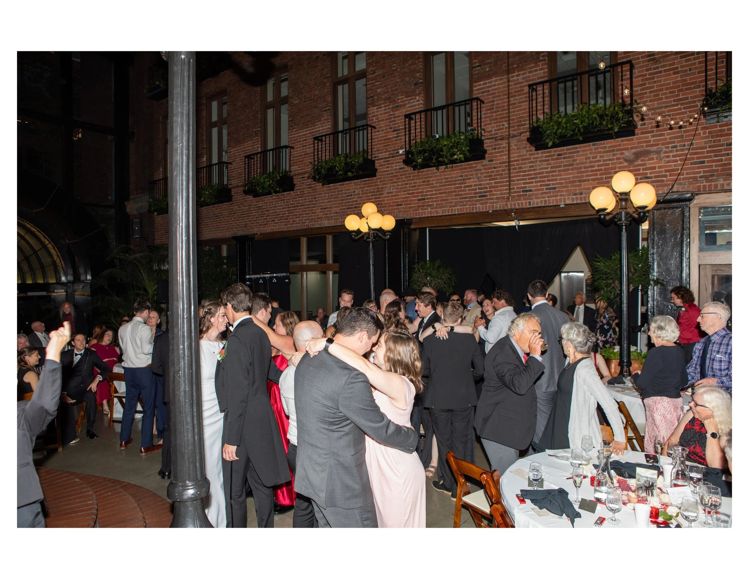 People dancing closely at a wedding reception in a rustic indoor venue with brick walls, string lights, and elegant decor.