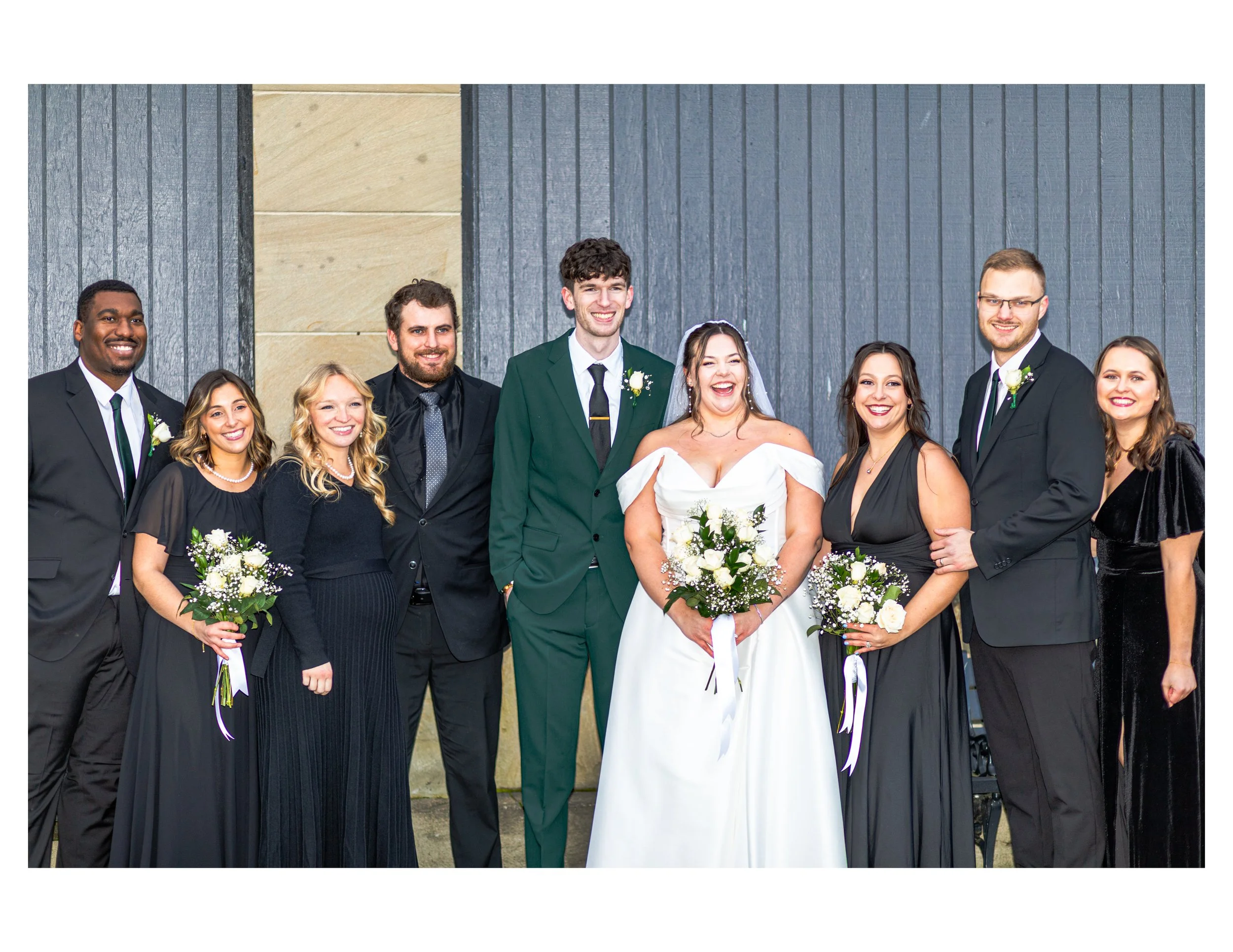 A group of smiling people dressed in formal attire, including a bride in a white dress holding a bouquet and a groom in a green suit, posing outdoors against a gray wooden wall.
