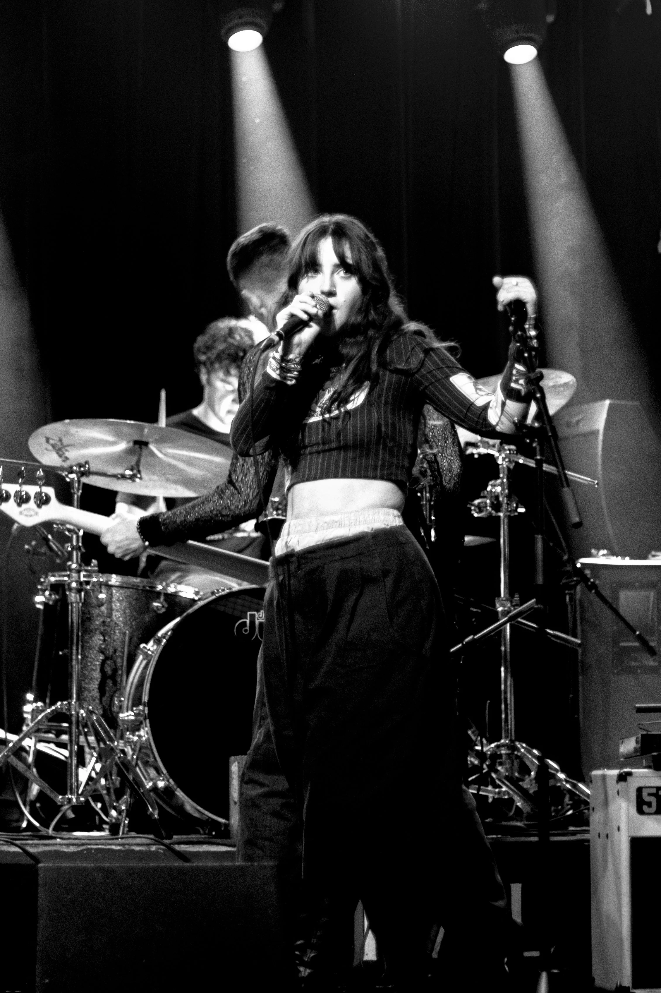 A woman singing into a microphone on stage with a band playing behind her, under stage lights.