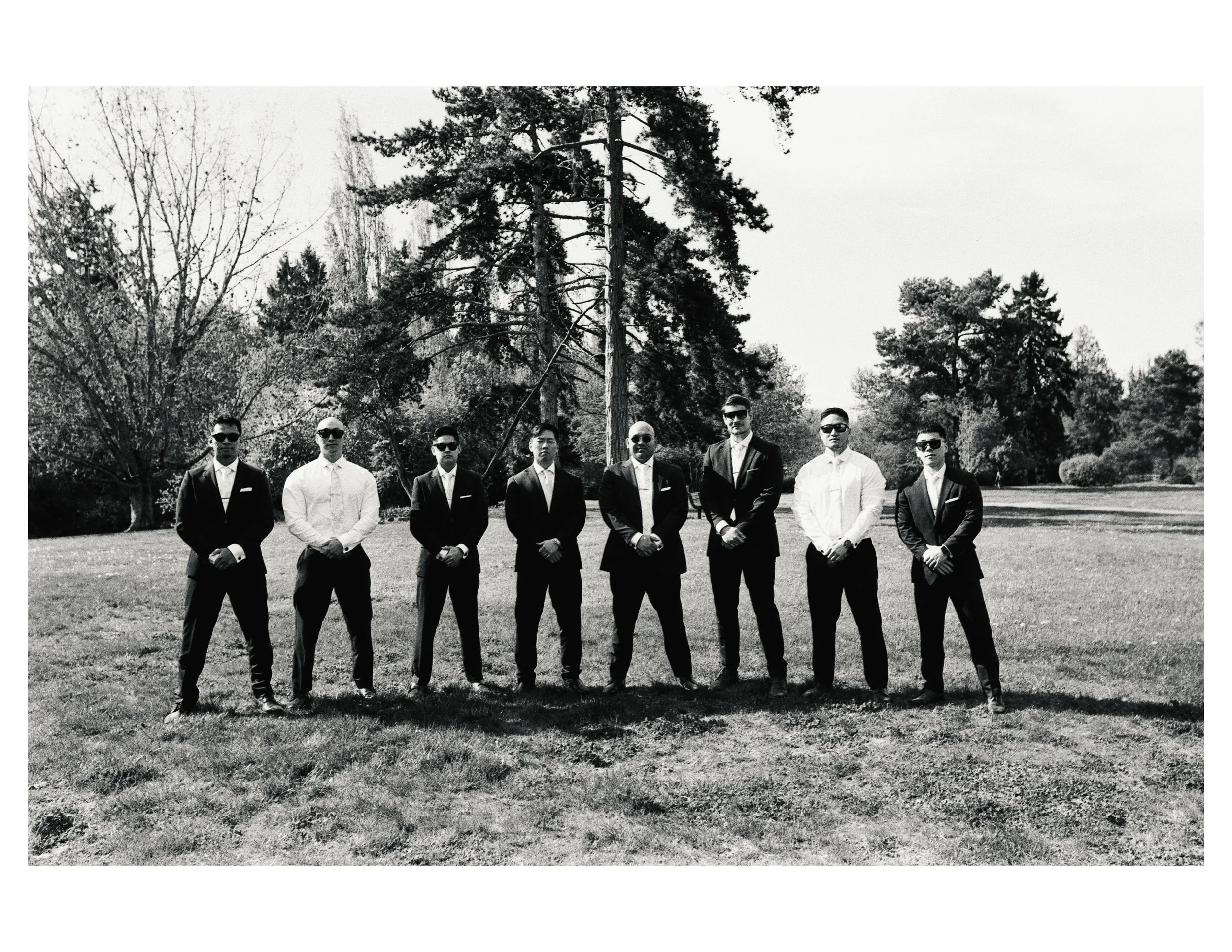 A black and white photo of nine men standing outdoors in a line, dressed in formal suits, with trees in the background.