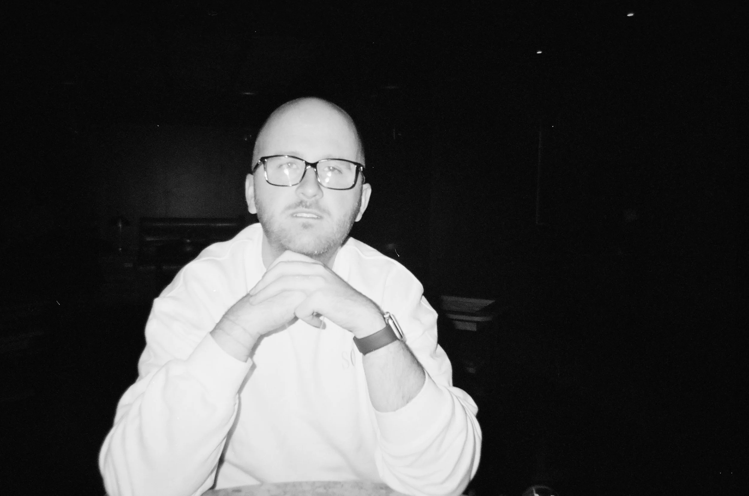 A man with glasses and a beard, wearing a white shirt, is sitting at a table with his hands clasped in front of him, looking at the camera in a dimly lit environment.