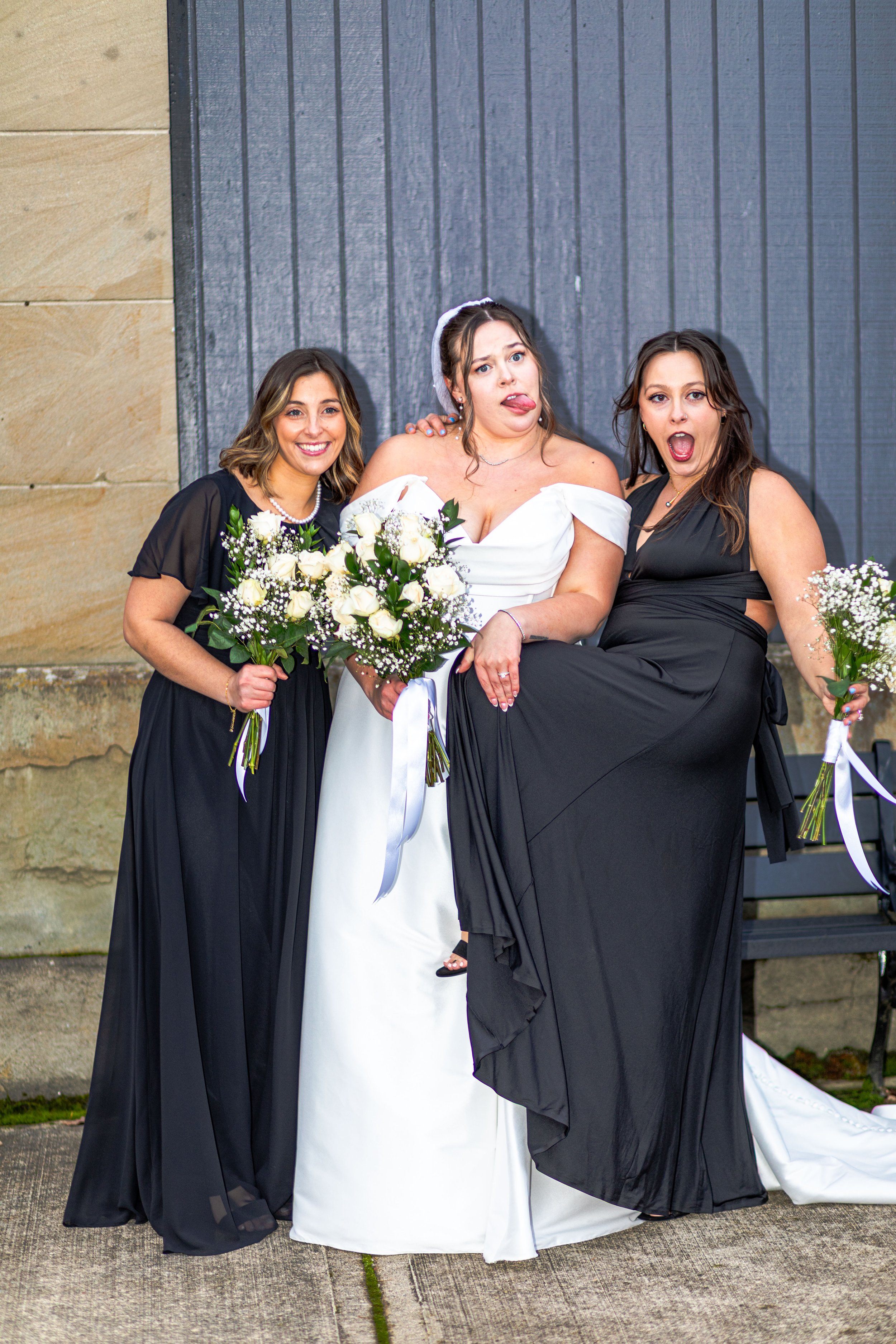 Three women at a wedding, with the bride in a white gown and two bridesmaids in black dresses, all holding bouquets of white flowers, standing against a wooden wall.