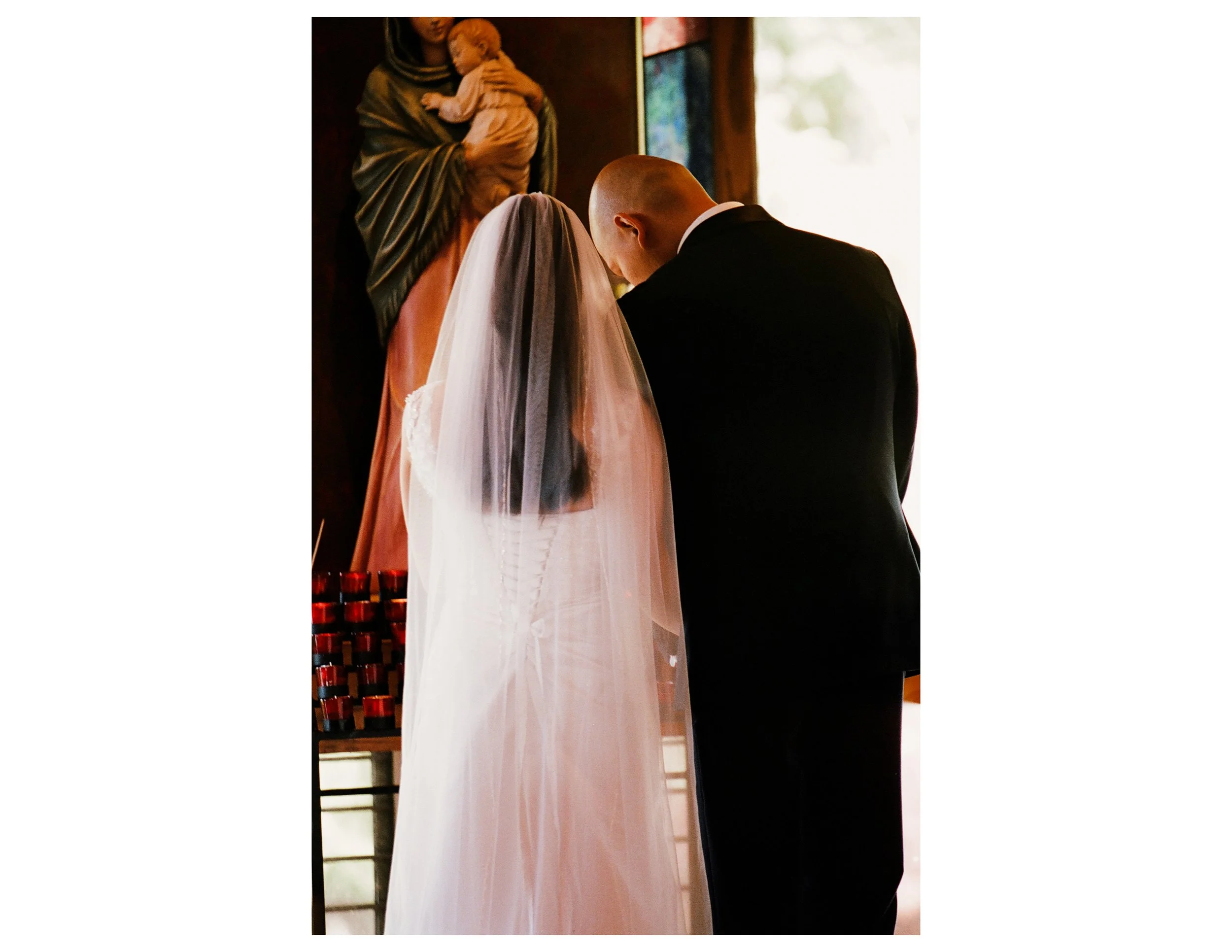A couple appears to be kneeling at the altar in a church, possibly during a wedding ceremony. The woman is wearing a white wedding gown and veil, while the man is dressed in a black suit and white shirt. In the background, there's a religious figure 